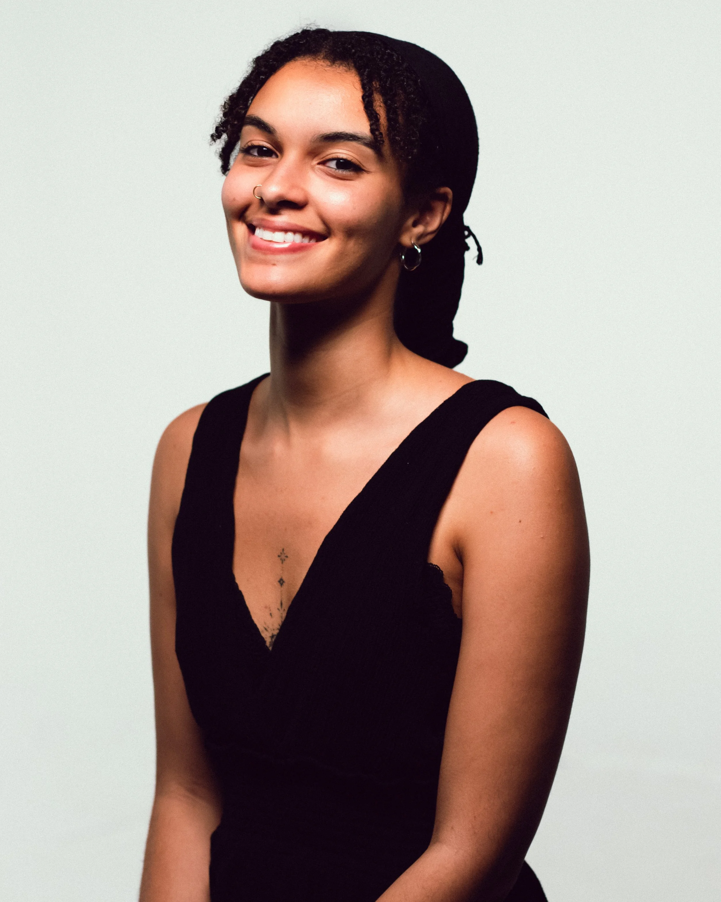 Portrait of a smiling young woman with curly hair, wearing a black sleeveless top, against a plain light background.