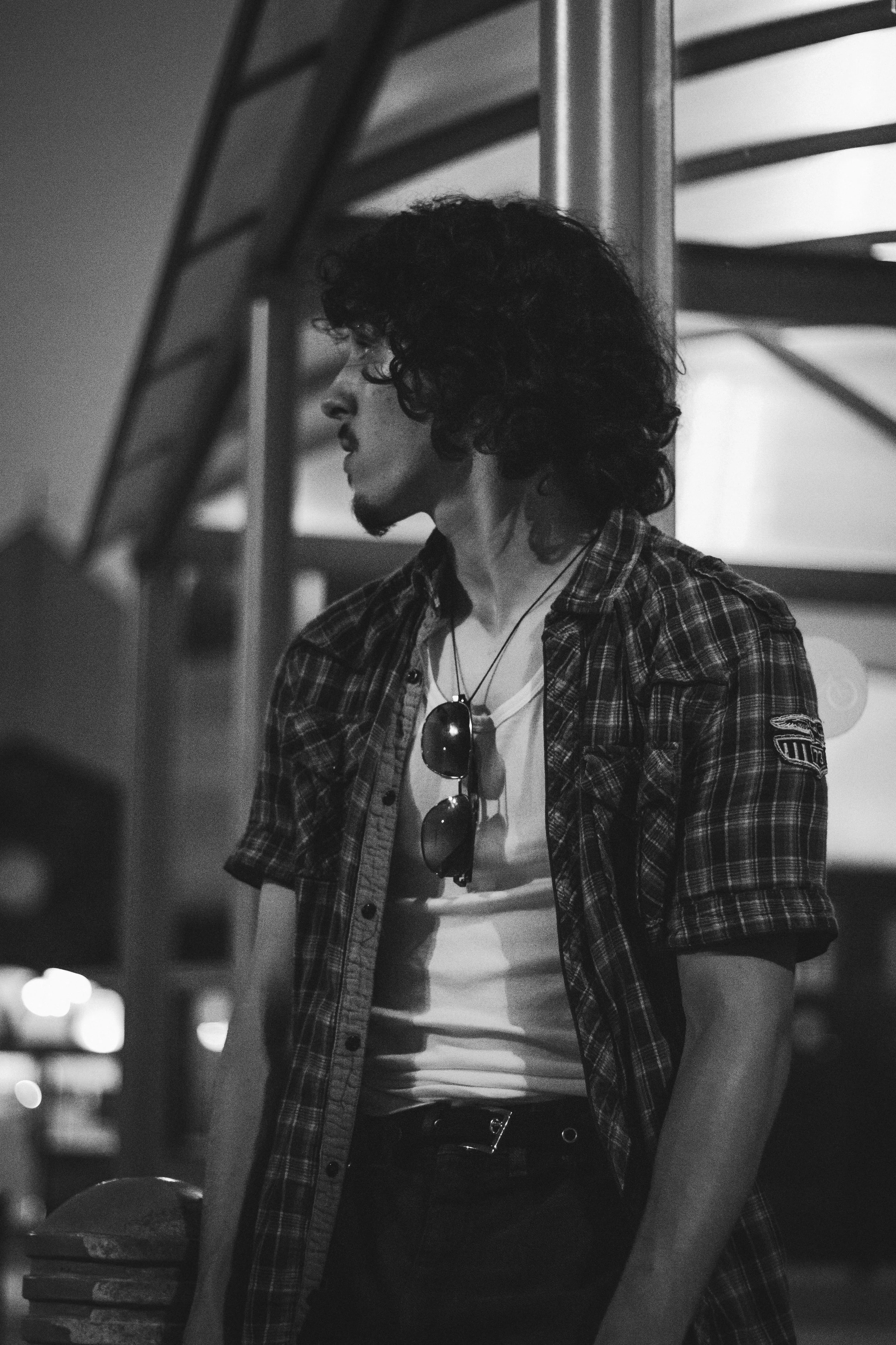 Black and white photo of a young person with curly hair wearing a plaid shirt, sunglasses hanging around their neck, t-shirt, and jeans, standing next to a pole at night.