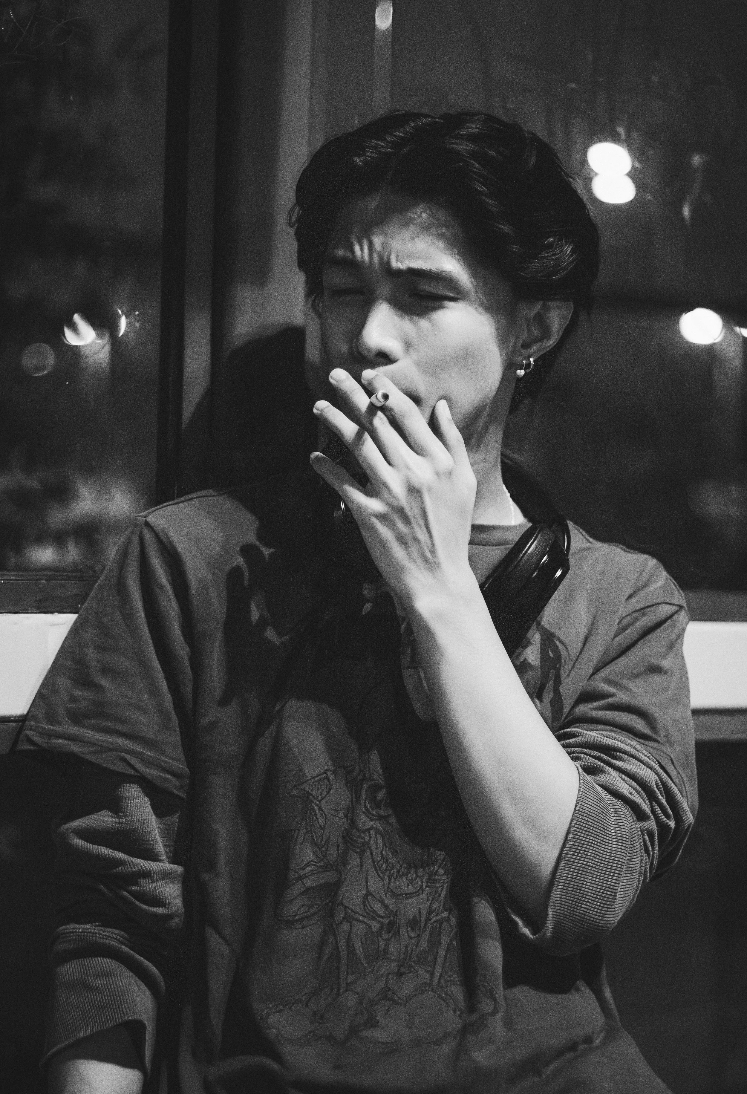 A person with styled hair, earrings, and a graphic t-shirt, smoking a cigarette in a dimly lit room with a reflective window and blurred lights outside.