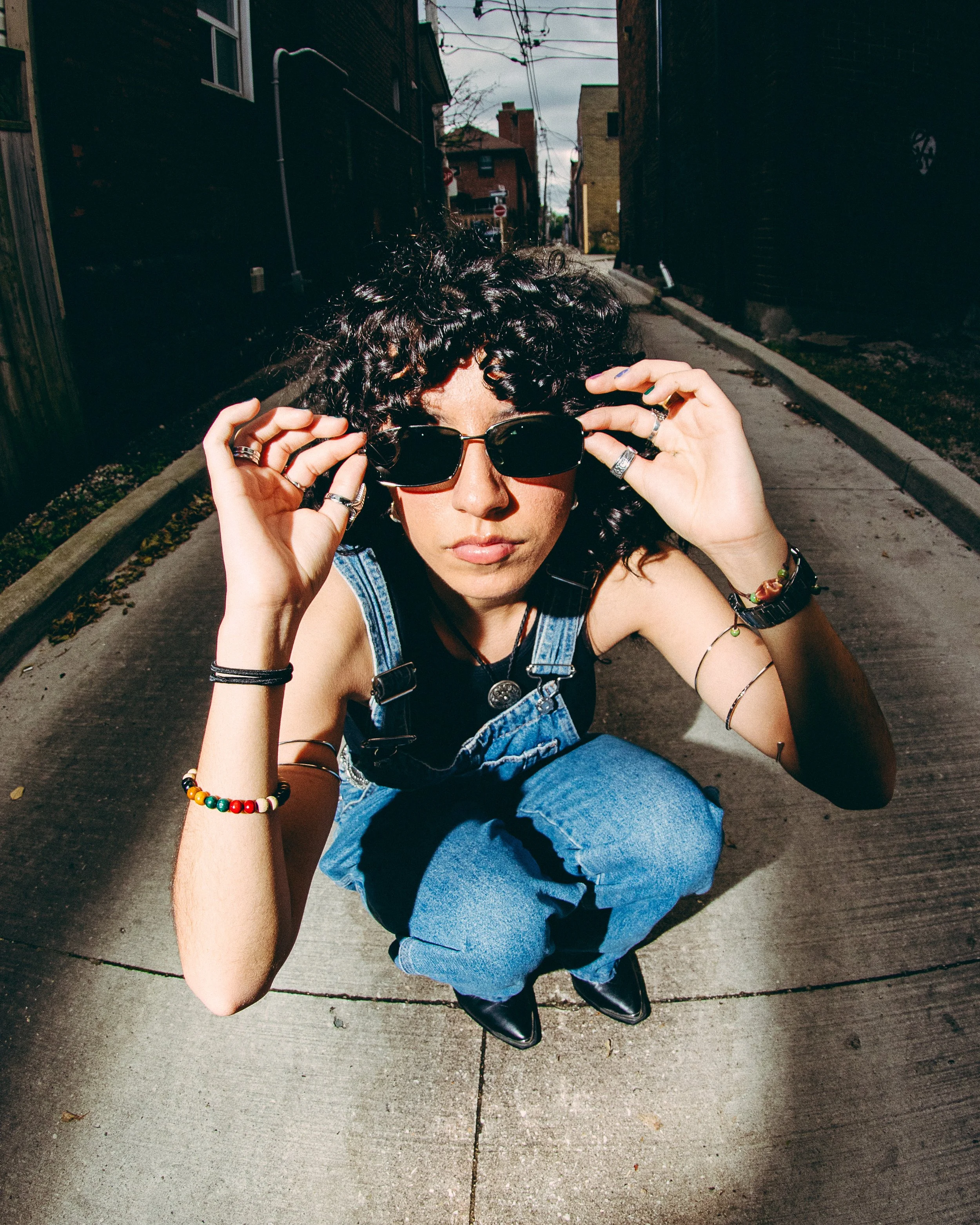 A woman with curly black hair wearing sunglasses, a black sleeveless top, and denim overalls crouching on a sidewalk in an urban alley, adjusting her sunglasses.