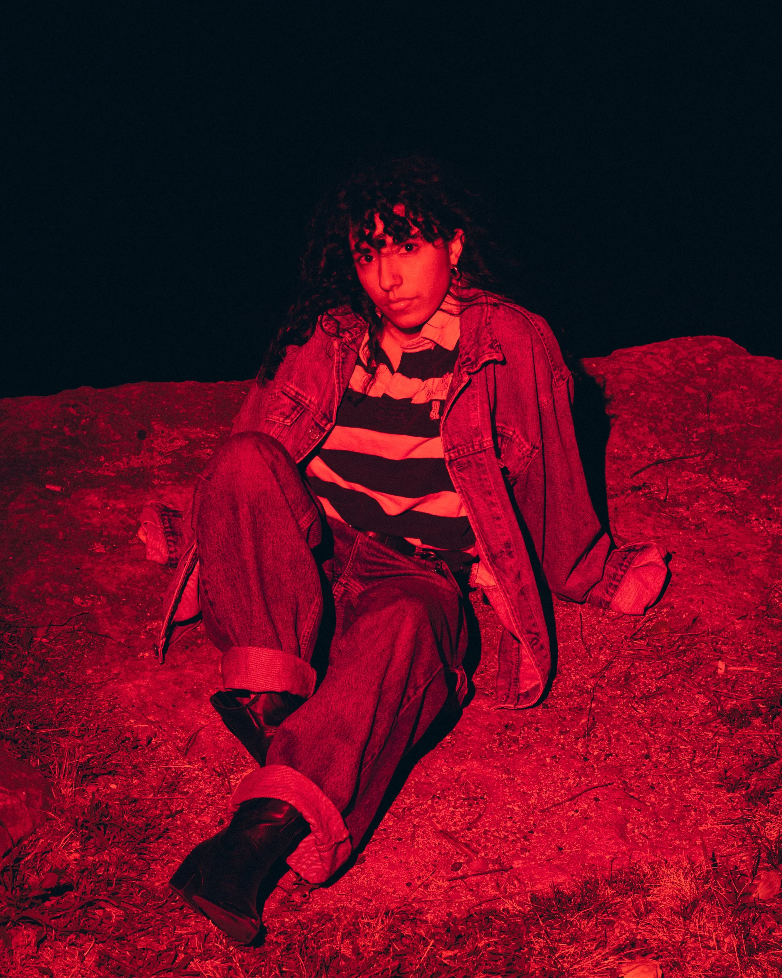 Person sitting on ground at night illuminated by red light, wearing striped shirt, denim jacket, jeans, and boots.