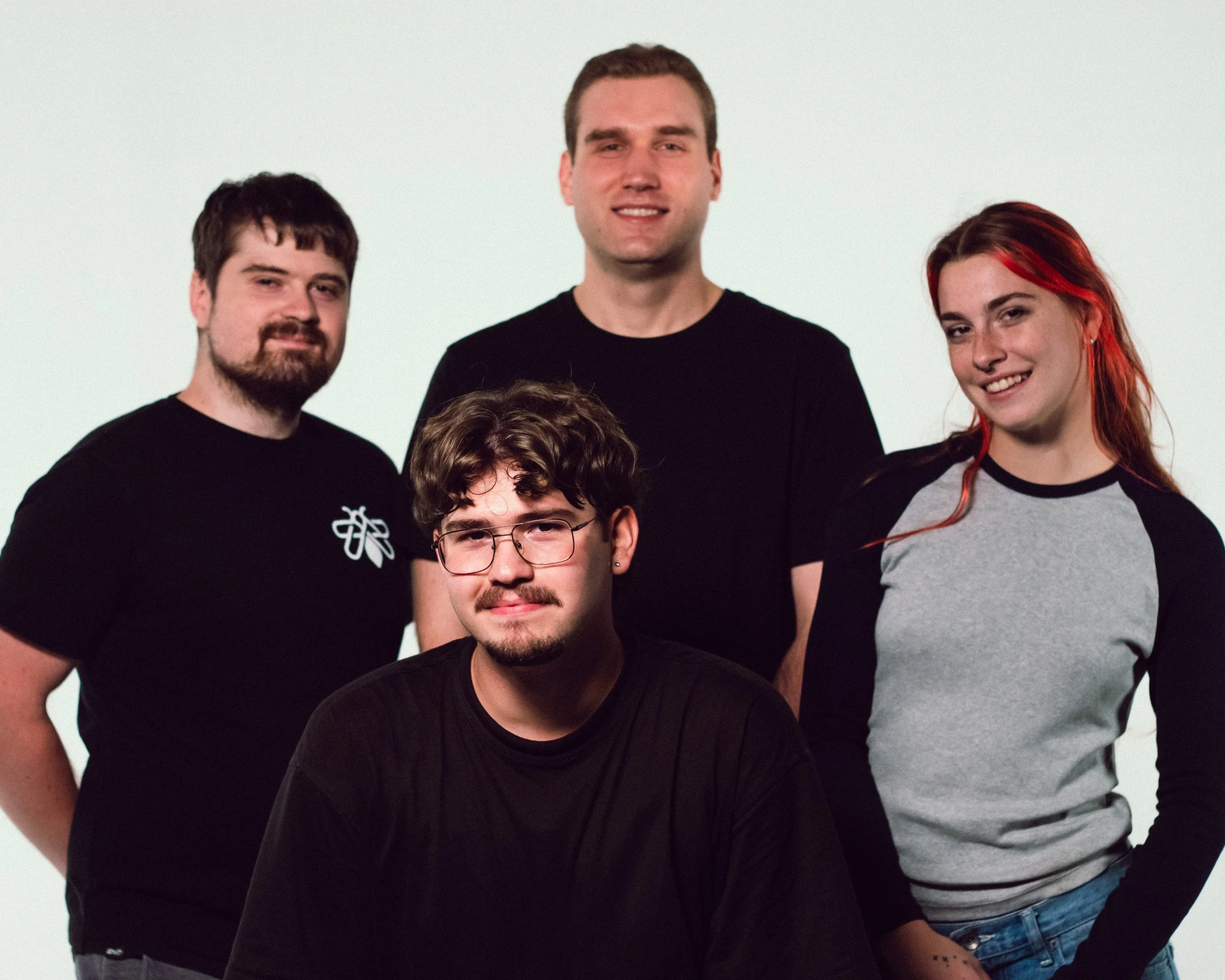 Group portrait of four young adults, three men and one woman, standing against a plain white background. The man in front has curly hair, glasses, and a beard, wearing a black shirt. The woman on the right has long red hair and is wearing a grey and 
