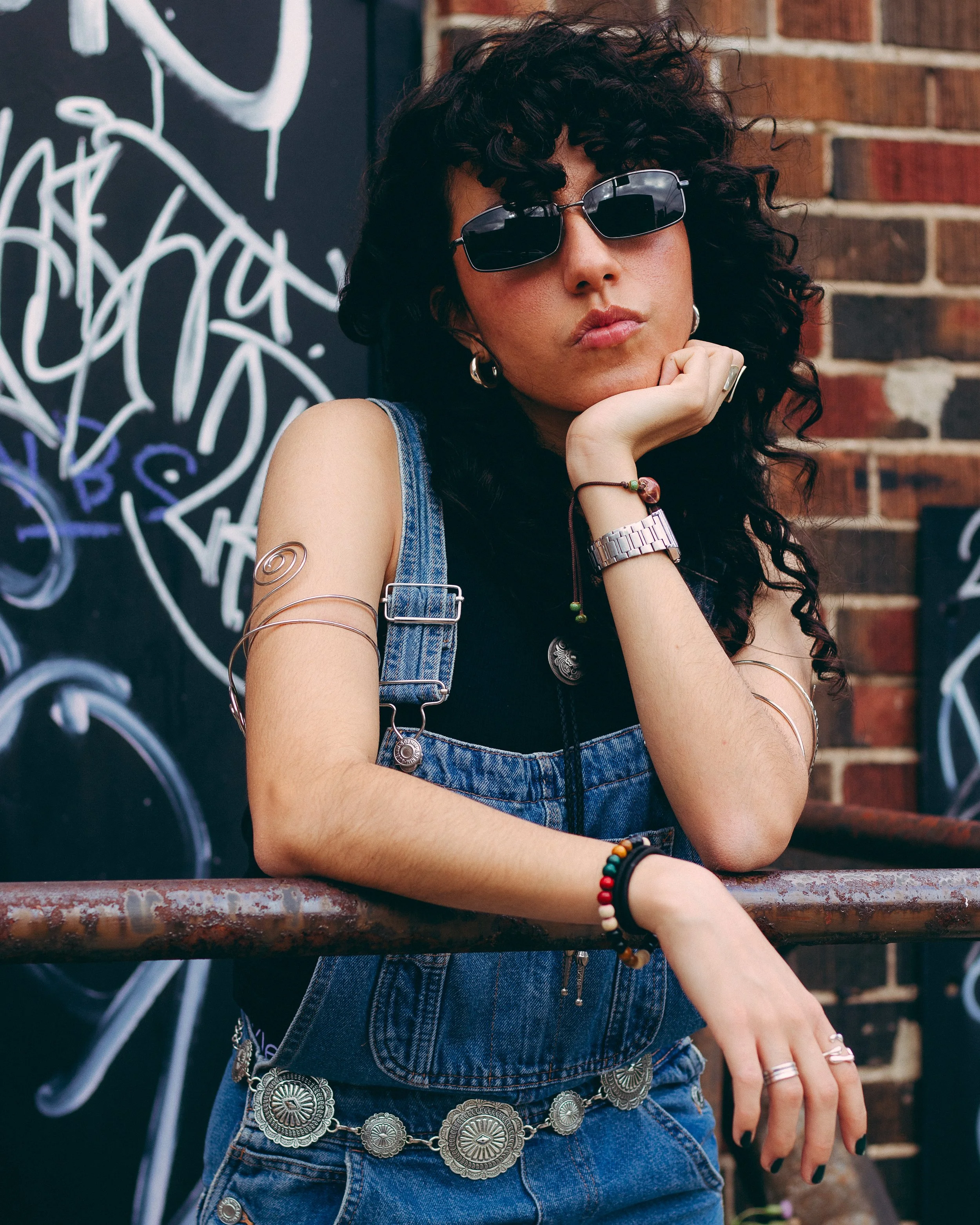 A young woman with curly dark hair and sunglasses leaning on a rusty railing in front of a graffiti-covered wall and brick building.