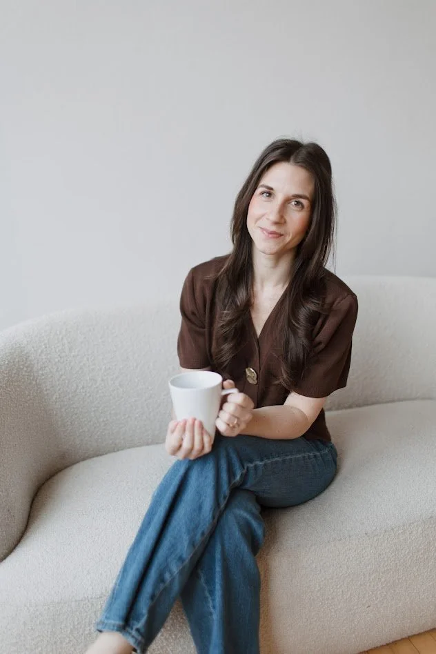 A woman with long dark hair, wearing a brown shirt, sitting on a light-colored sofa, holding a white mug, and smiling at the camera.