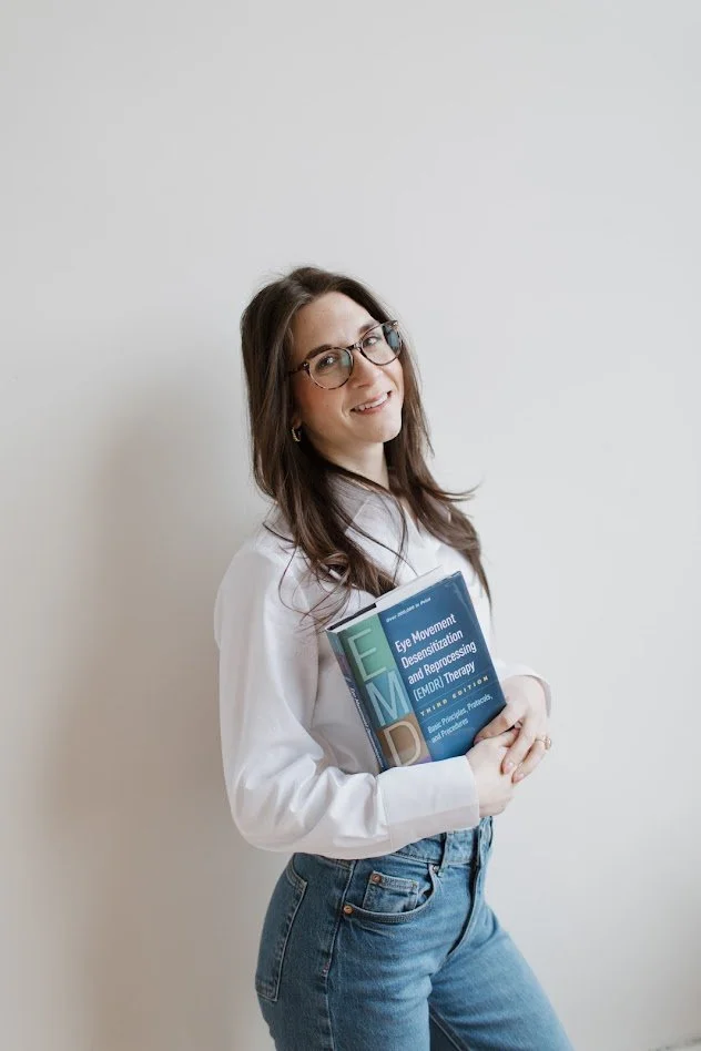A woman with glasses and long brown hair holding a book titled 'Eye Movement Desensitization and Reprocessing (EMDR) Therapy' against a plain white wall.