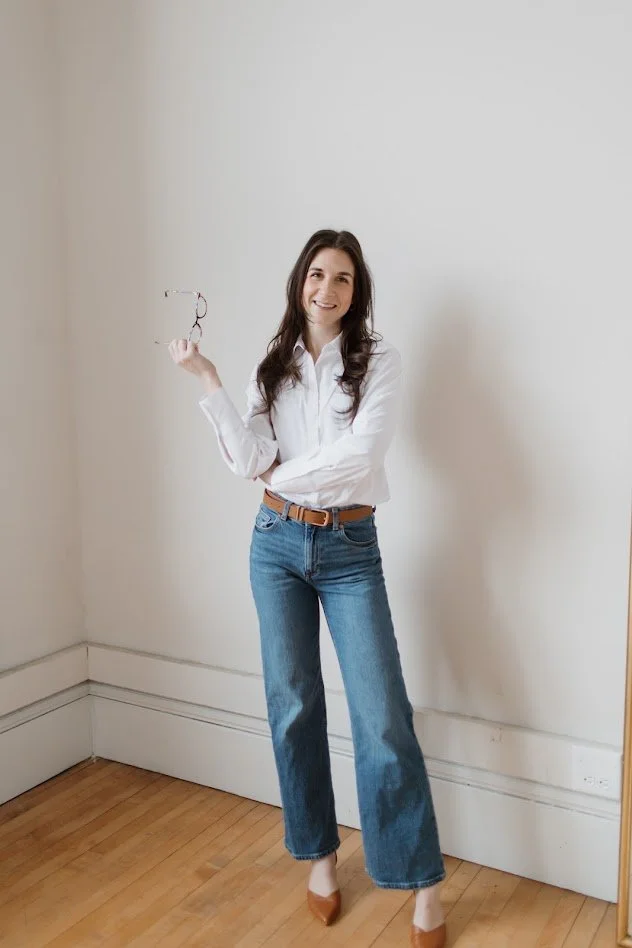 A woman with long dark hair standing in a room with white walls and wooden floor, smiling and holding a pair of glasses in her right hand.