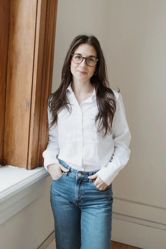A young woman with long dark hair, glasses, and hoop earrings stands indoors near a wooden window frame, wearing a white button-up shirt and blue jeans, smiling softly with hands in her pockets.