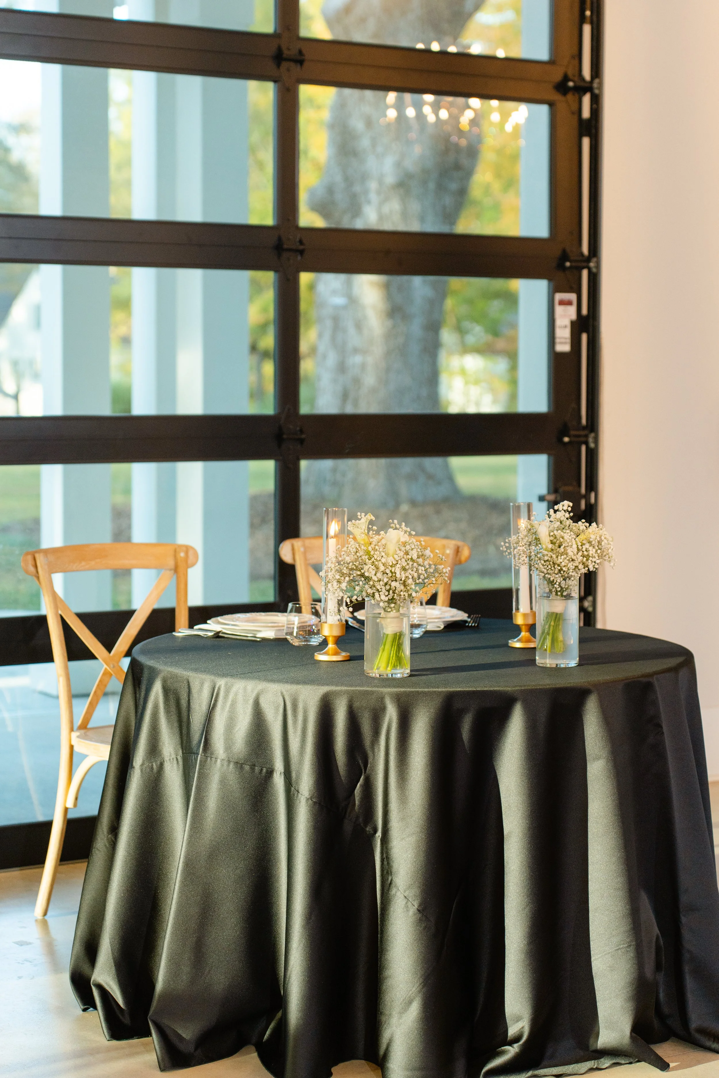 Round table with a black tablecloth decorated with small white floral centerpieces and tall glass candle holders, set for a meal, near a large window with a view of a tree outside.