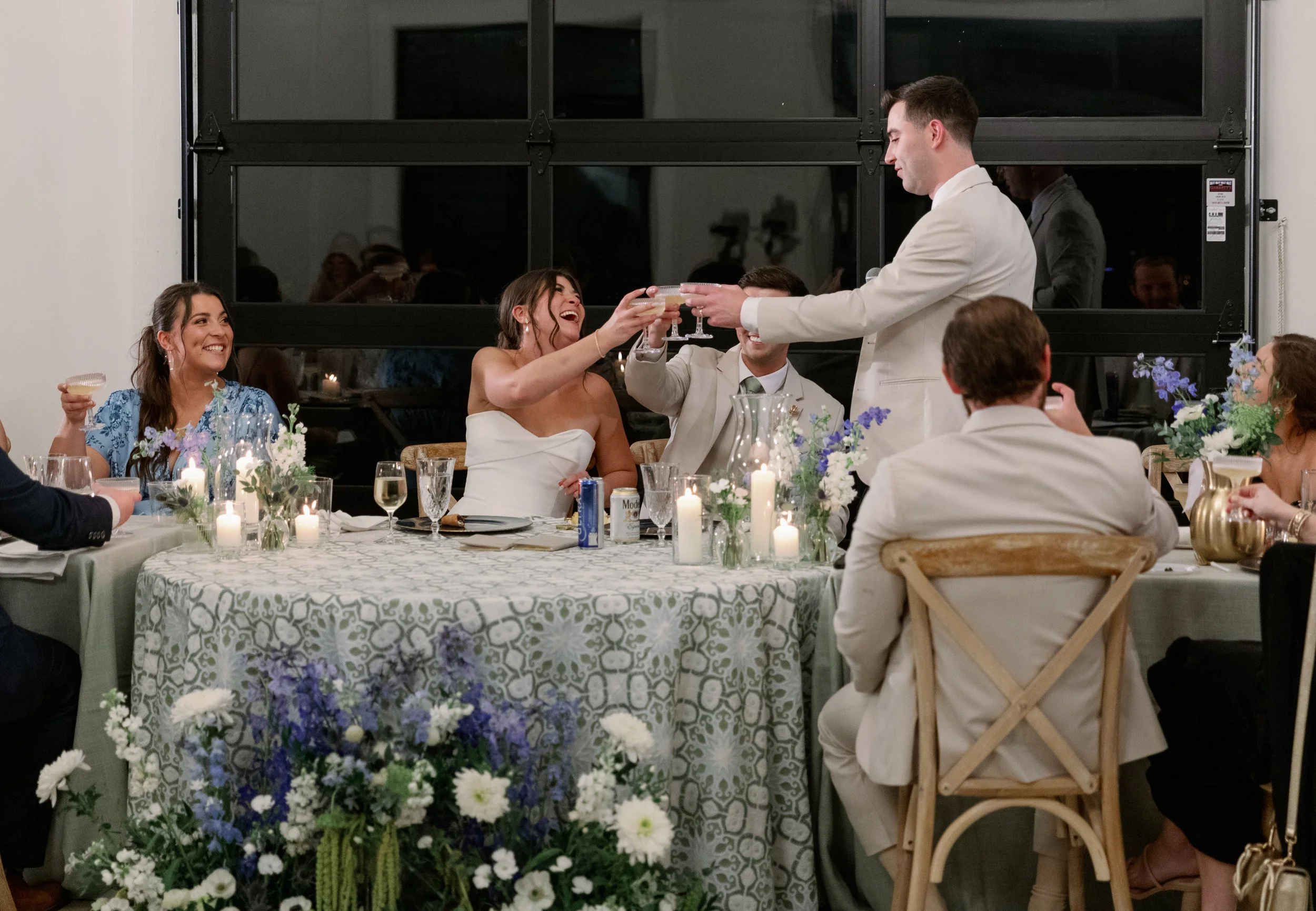 A wedding reception with a bride and groom toasting with guests at a decorated table with candles and flowers.