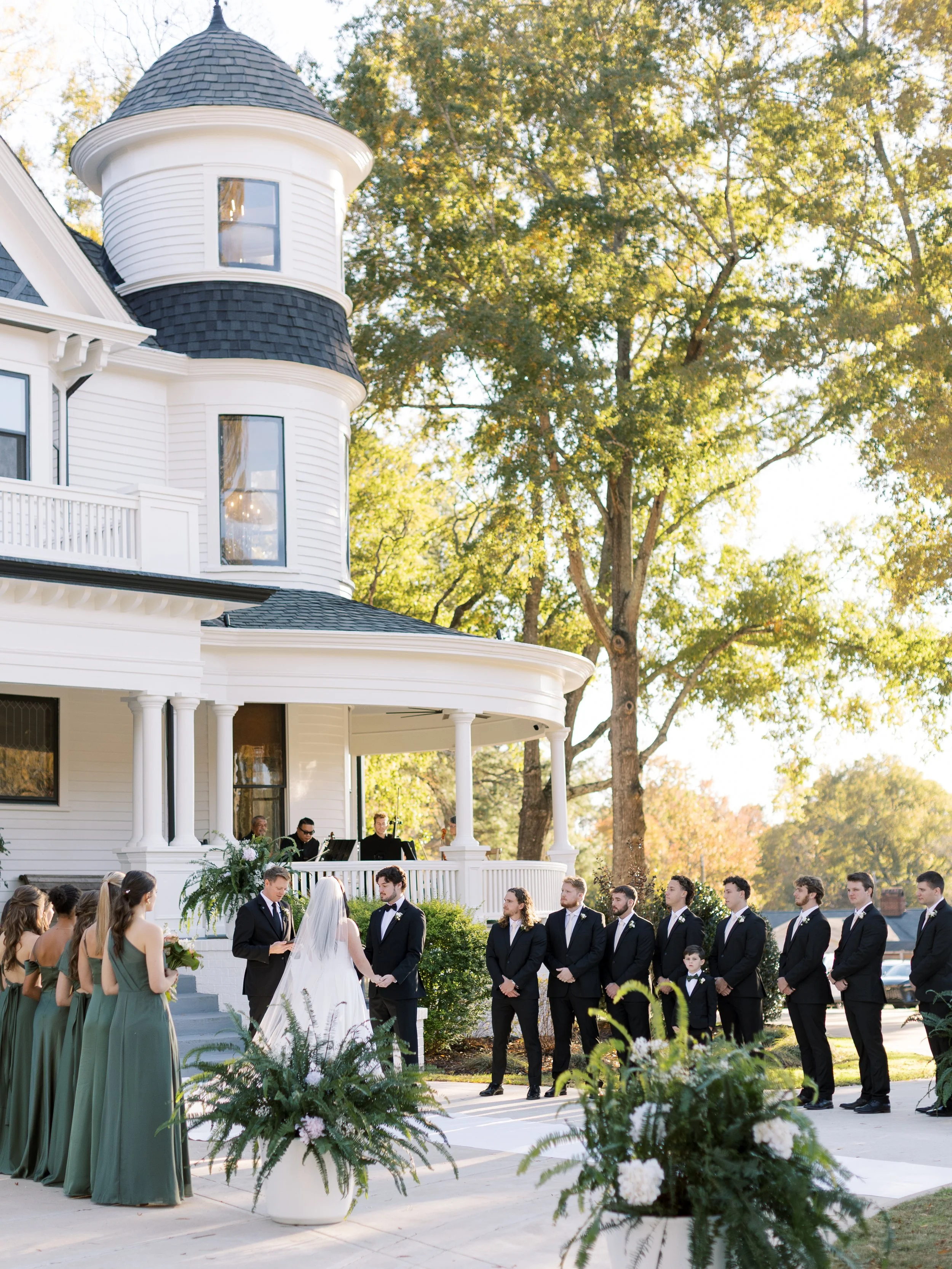 A wedding ceremony taking place outdoors in front of a large white house with a turret. The bride and groom are standing in front of an officiant, exchanging vows, with wedding party members and guests watching. The scene is set in a sunny, autumn la