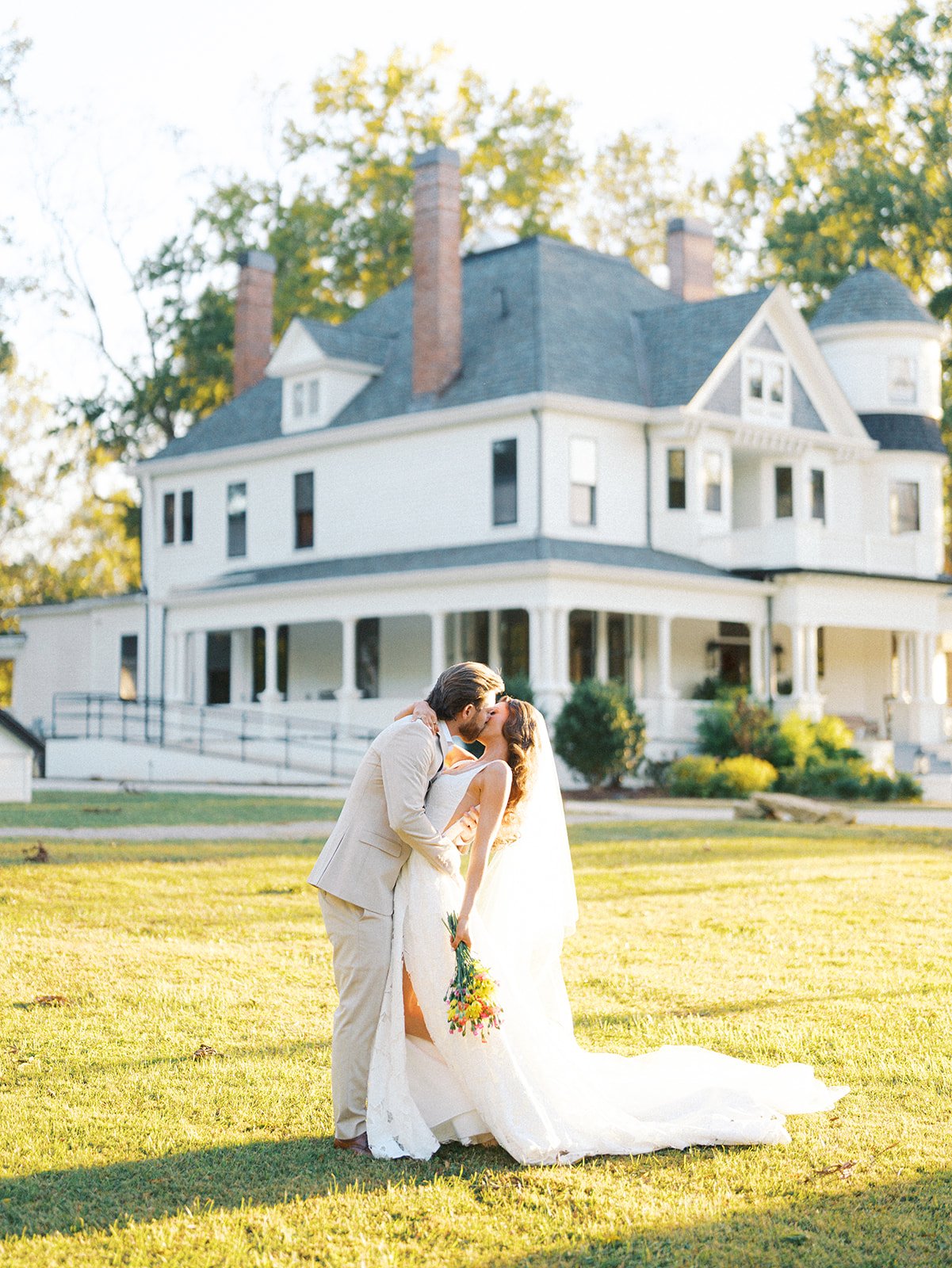 Bride and groom kissing outside in front of a large white house, holding a bouquet, during daylight.