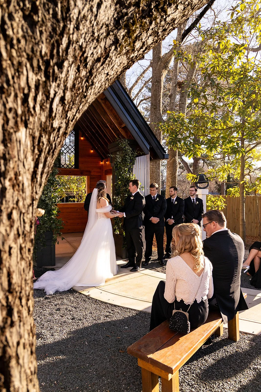 A wedding ceremony outdoors with a bride and groom exchanging vows, surrounded by four groomsmen and two guests seated on a wooden bench, in front of a small wooden building with trees and sunlight in the background.