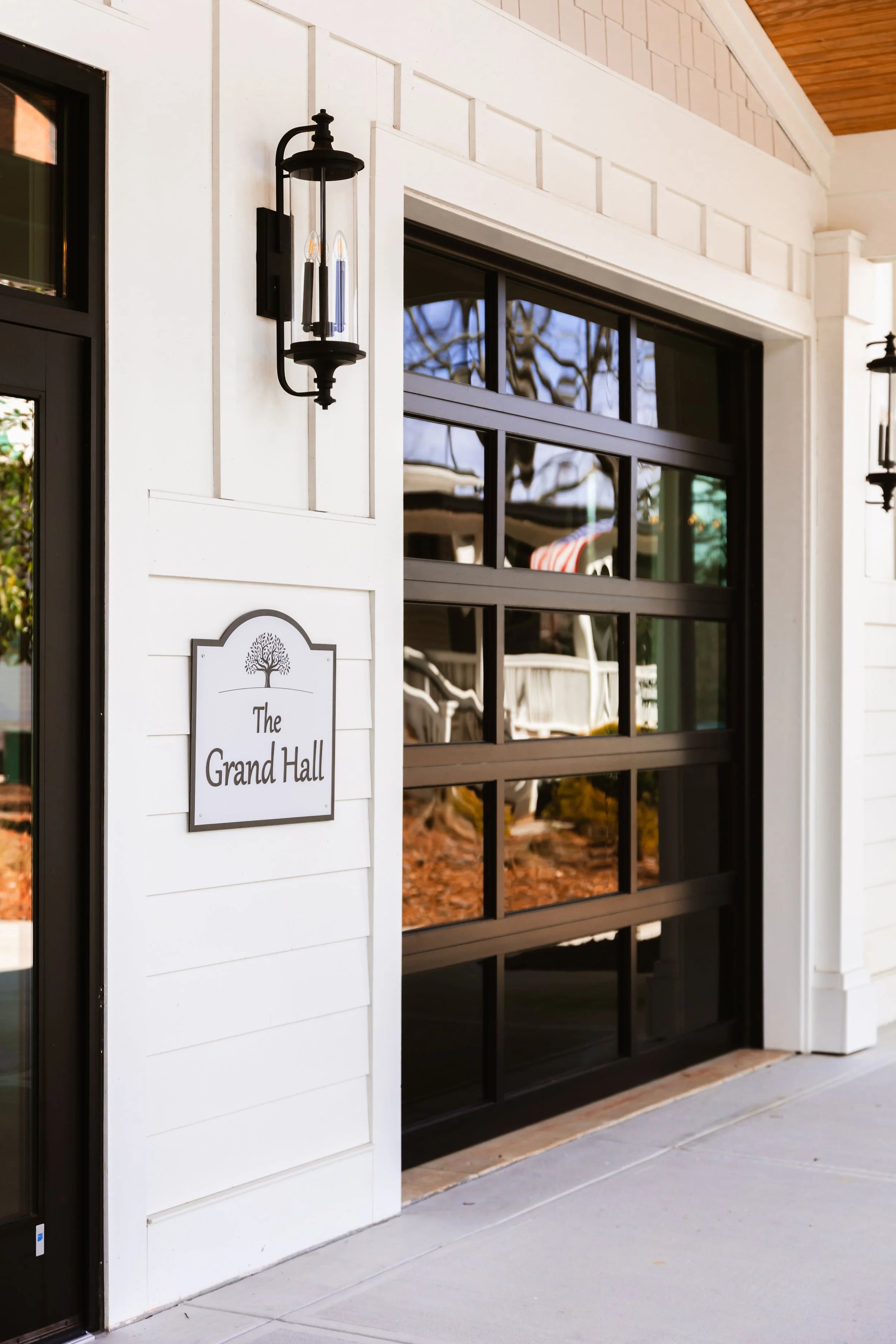 Exterior view of a house entrance with a black garage door, wall-mounted lantern, and a sign that reads "The Grand Hall" on a white wood-paneled wall.