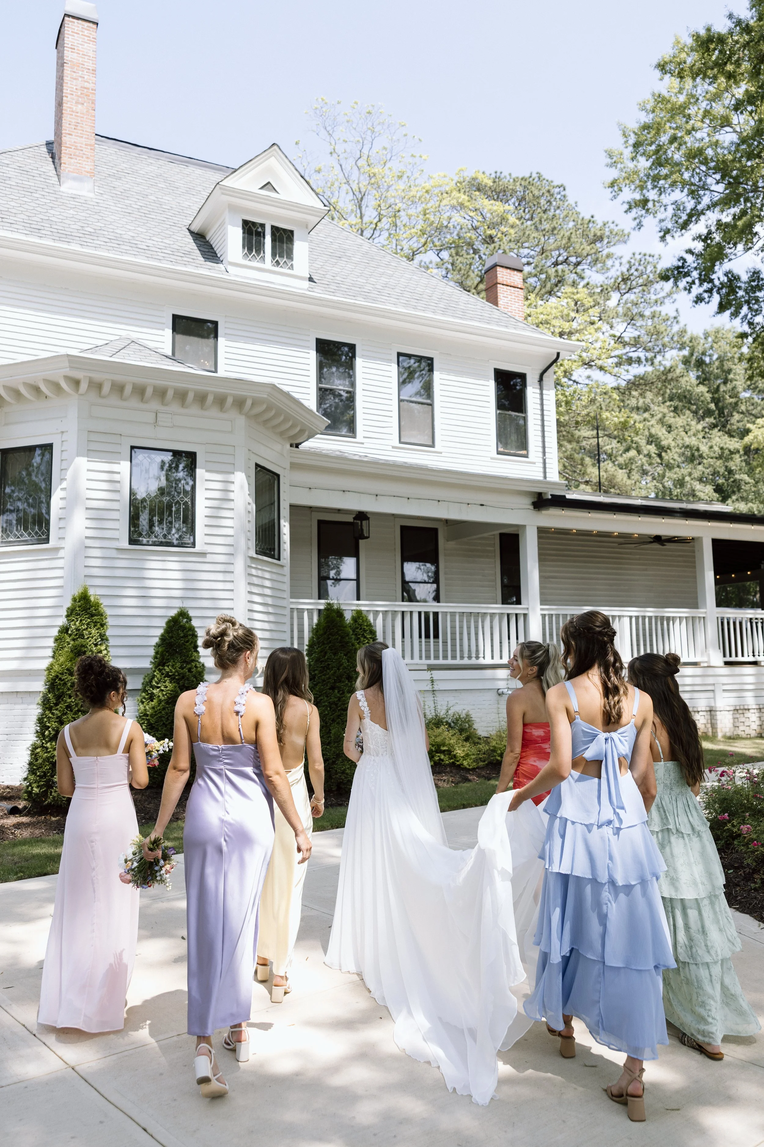 A bride in a white wedding gown walks with bridesmaids outside a white house with a porch, surrounded by trees.