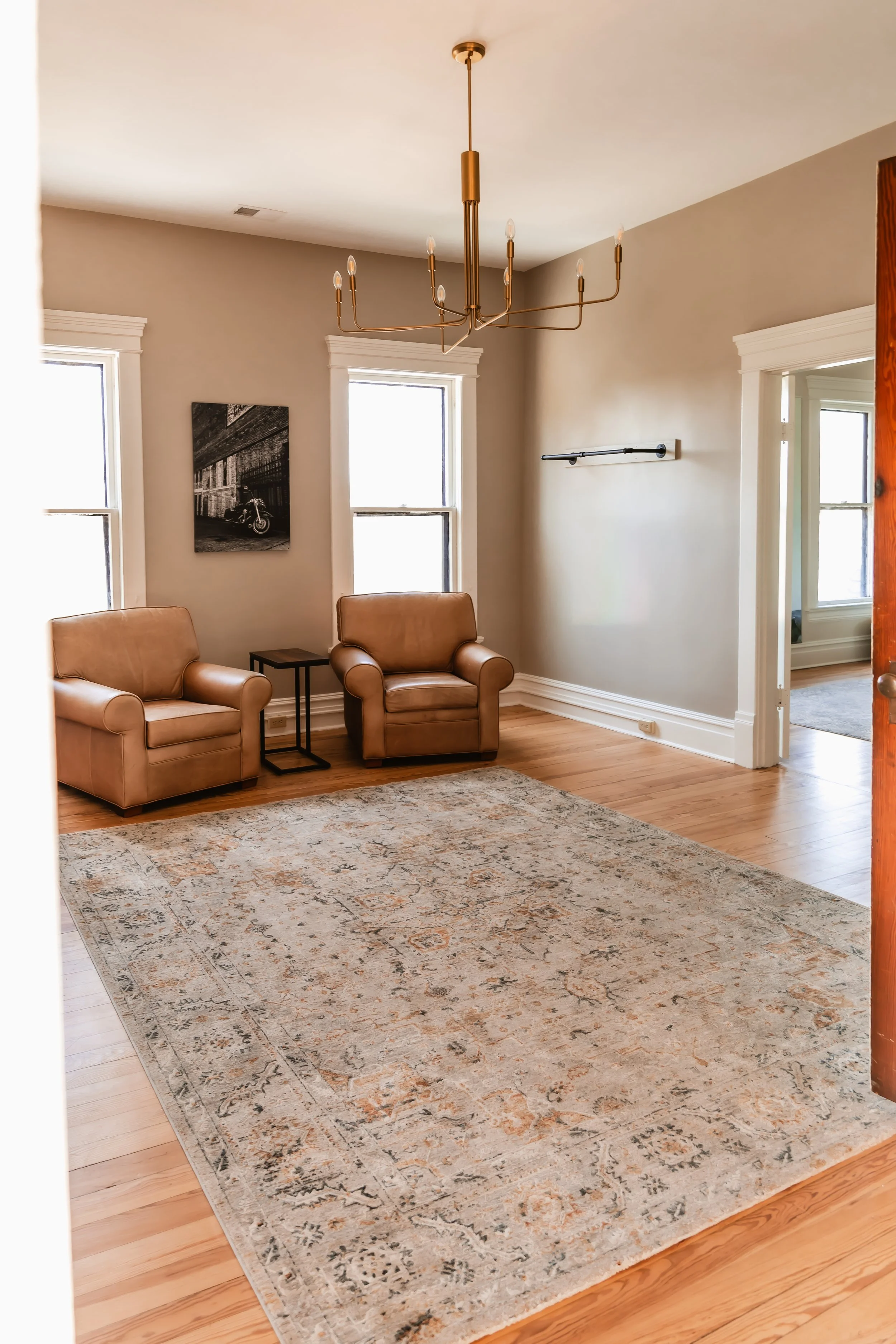 Living room with two tan leather armchairs, a patterned area rug, a small side table, two windows, and a modern chandelier.