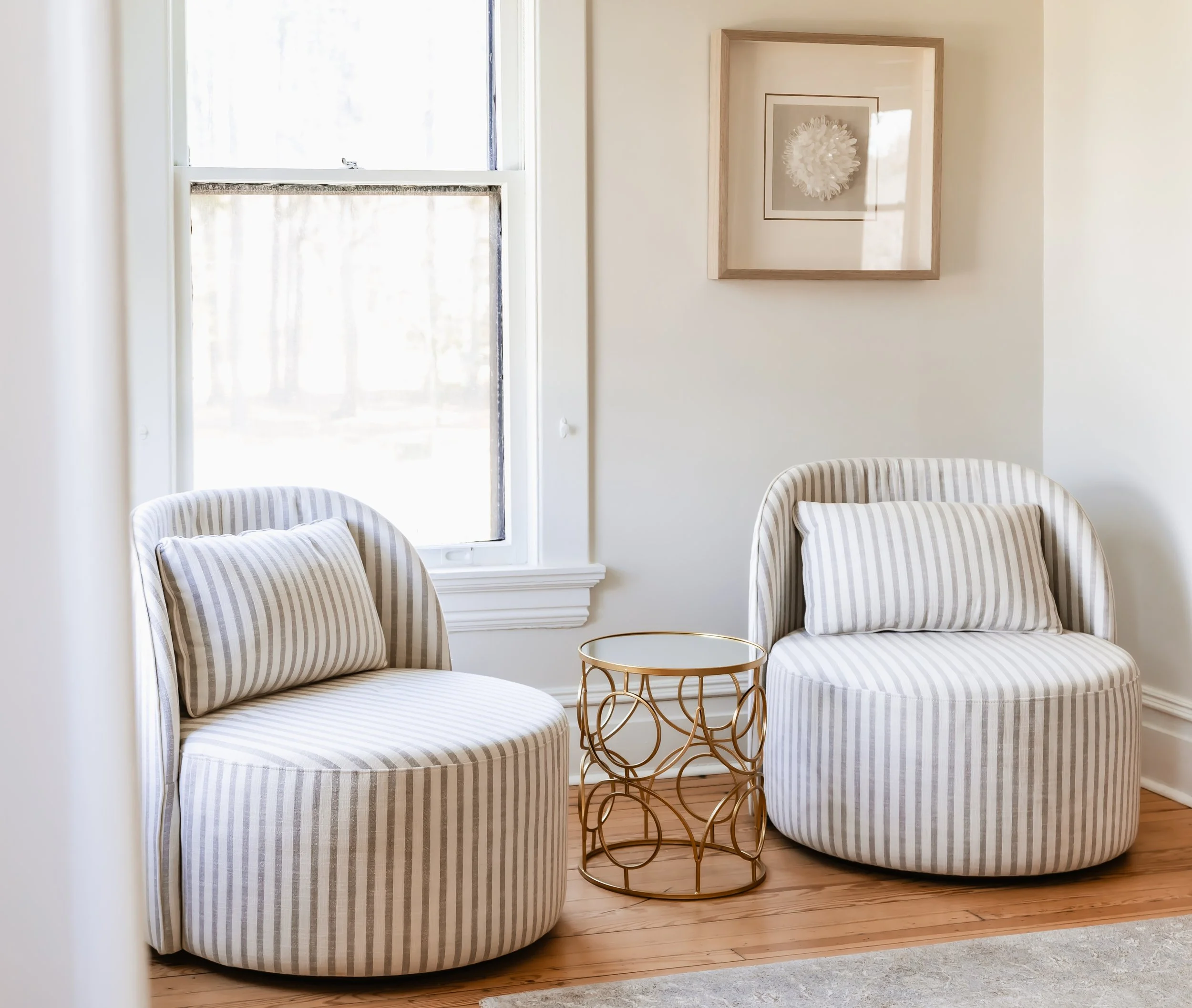 Two striped upholstered armchairs with matching pillows, a gold metal side table, and framed artwork on a cream wall near a window.