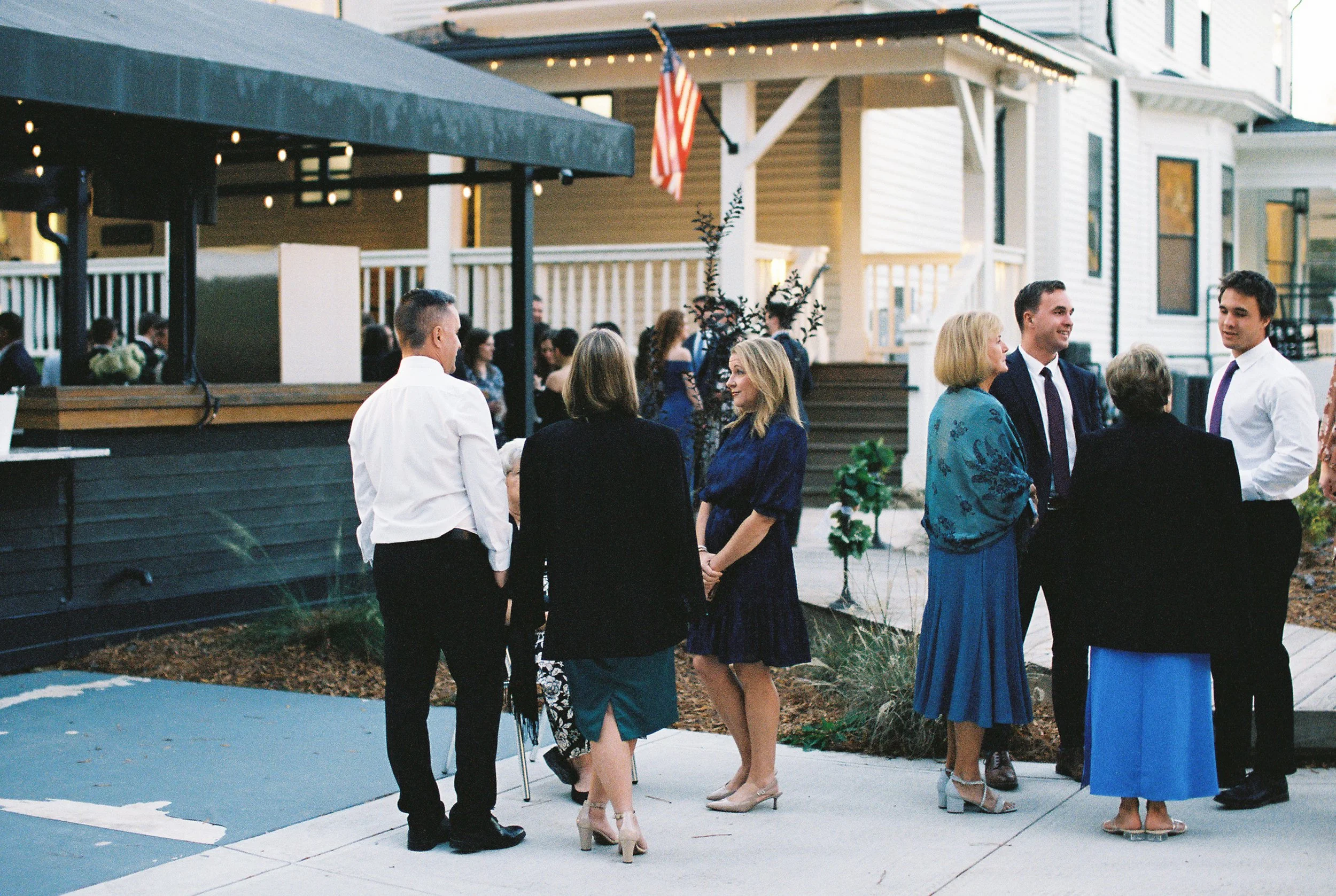 Group of people socializing outdoors at an event, standing near a building with stairs and an American flag, in the early evening.