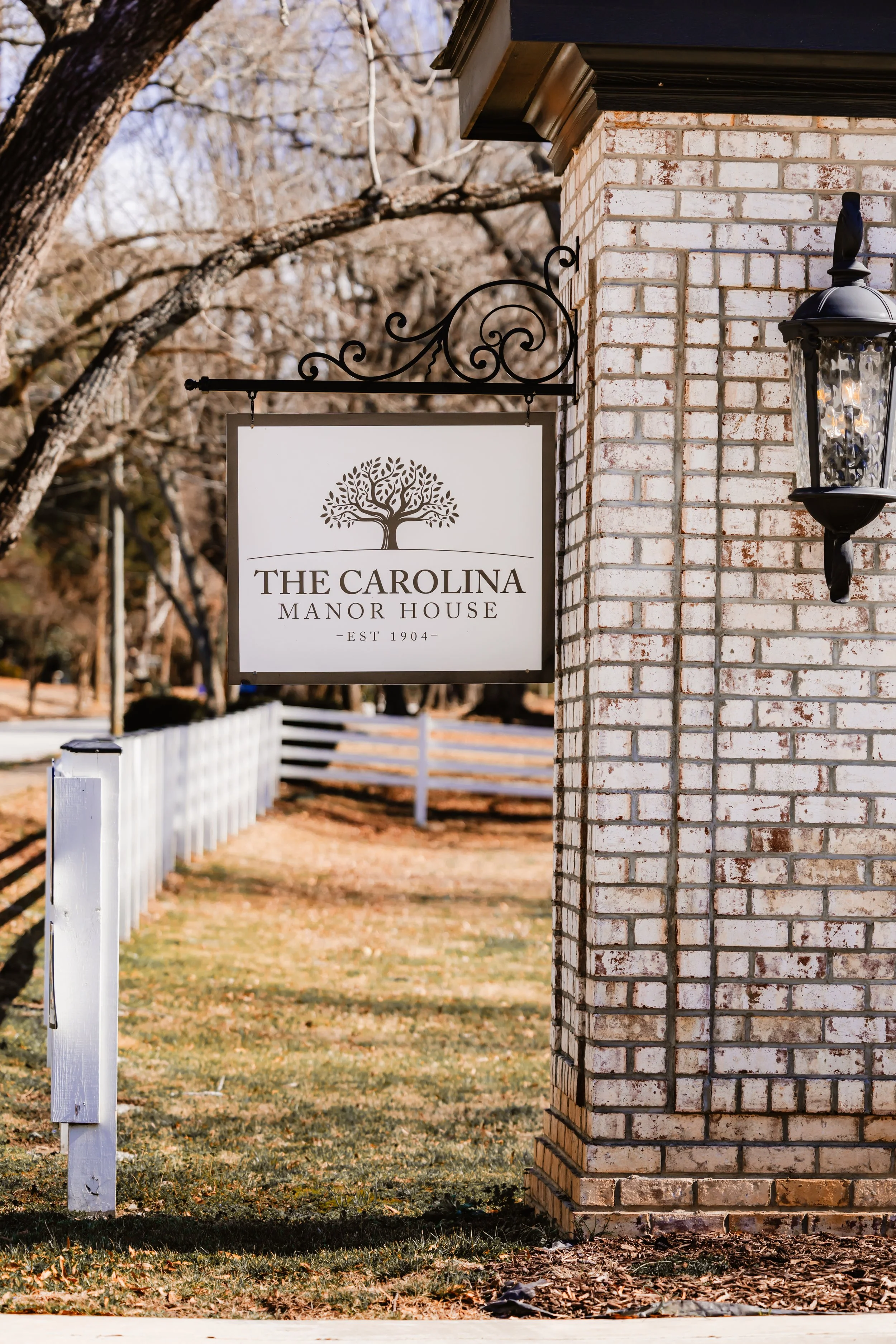 Sign for The Carolina Manor House with a tree logo, hanging on a decorative metal bracket attached to a brick wall, and a black outdoor lantern next to it.