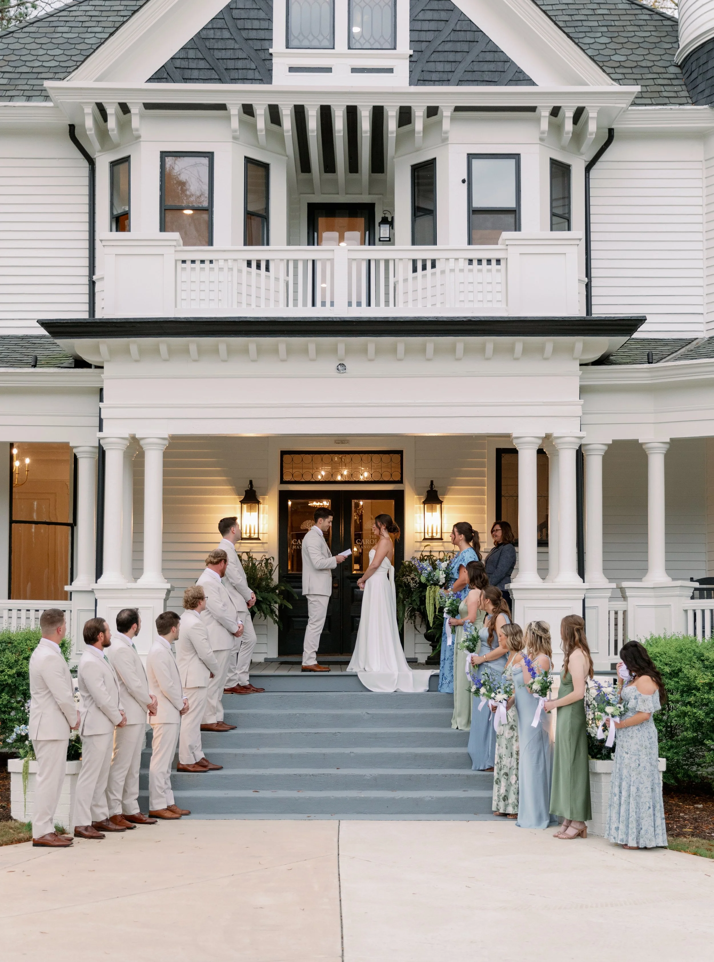 Wedding ceremony in front of a large white house, with the bride and groom standing on the steps. The bridal party and guests are gathered, some women holding bouquets, and all dressed in formal attire.