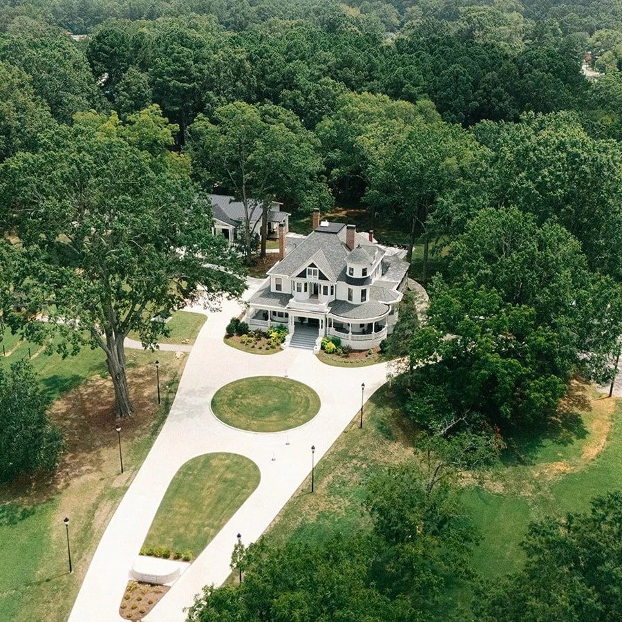 A large white Victorian-style house with a wrap-around porch, surrounded by green trees and a neatly maintained lawn. The driveway curves in front of the house, with lamp posts lining it, and there is a gravel area to the right.