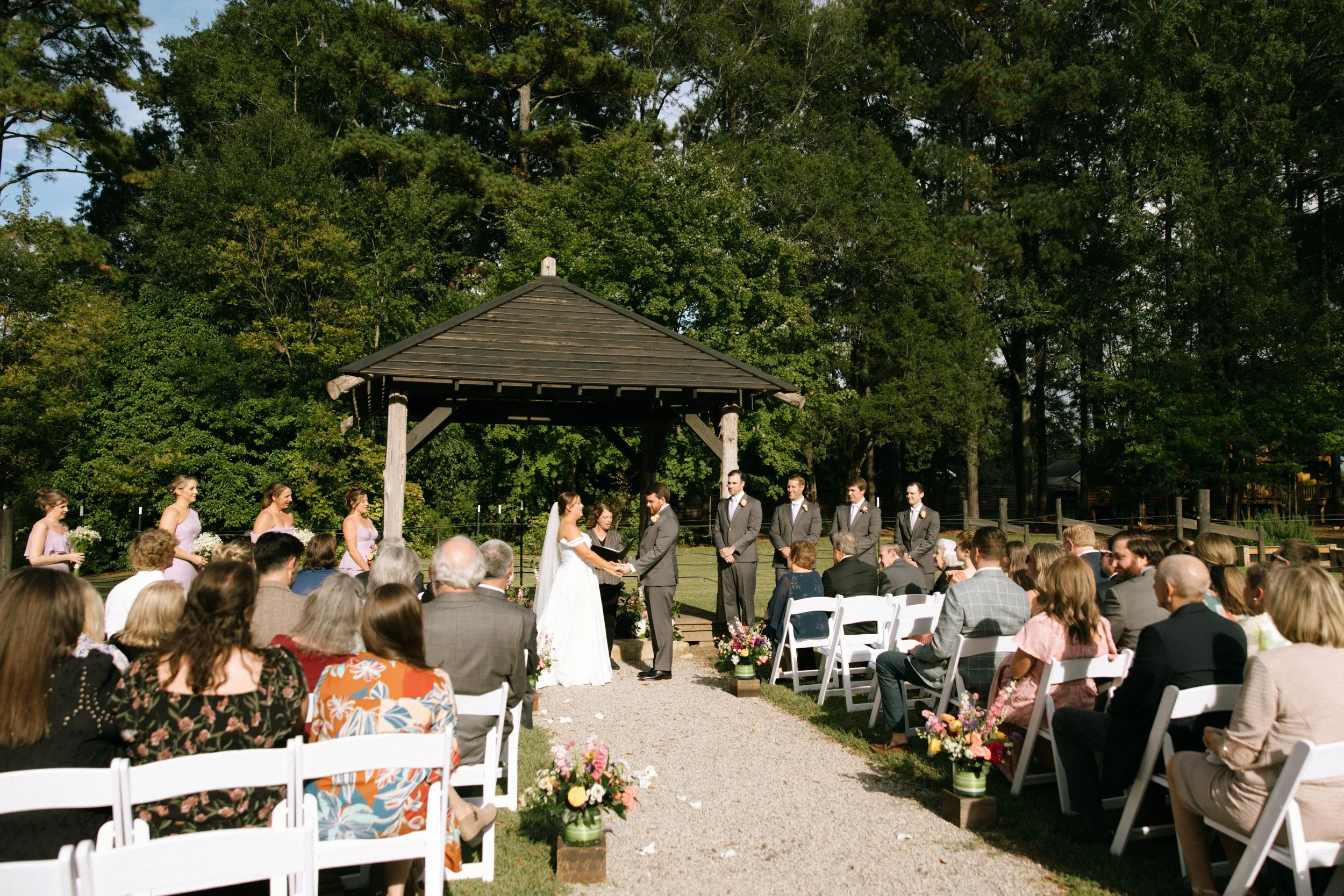 Outdoor wedding ceremony with a bride and groom exchanging vows under a pavilion, surrounded by bridesmaids and groomsmen, seated guests, and lush green trees.