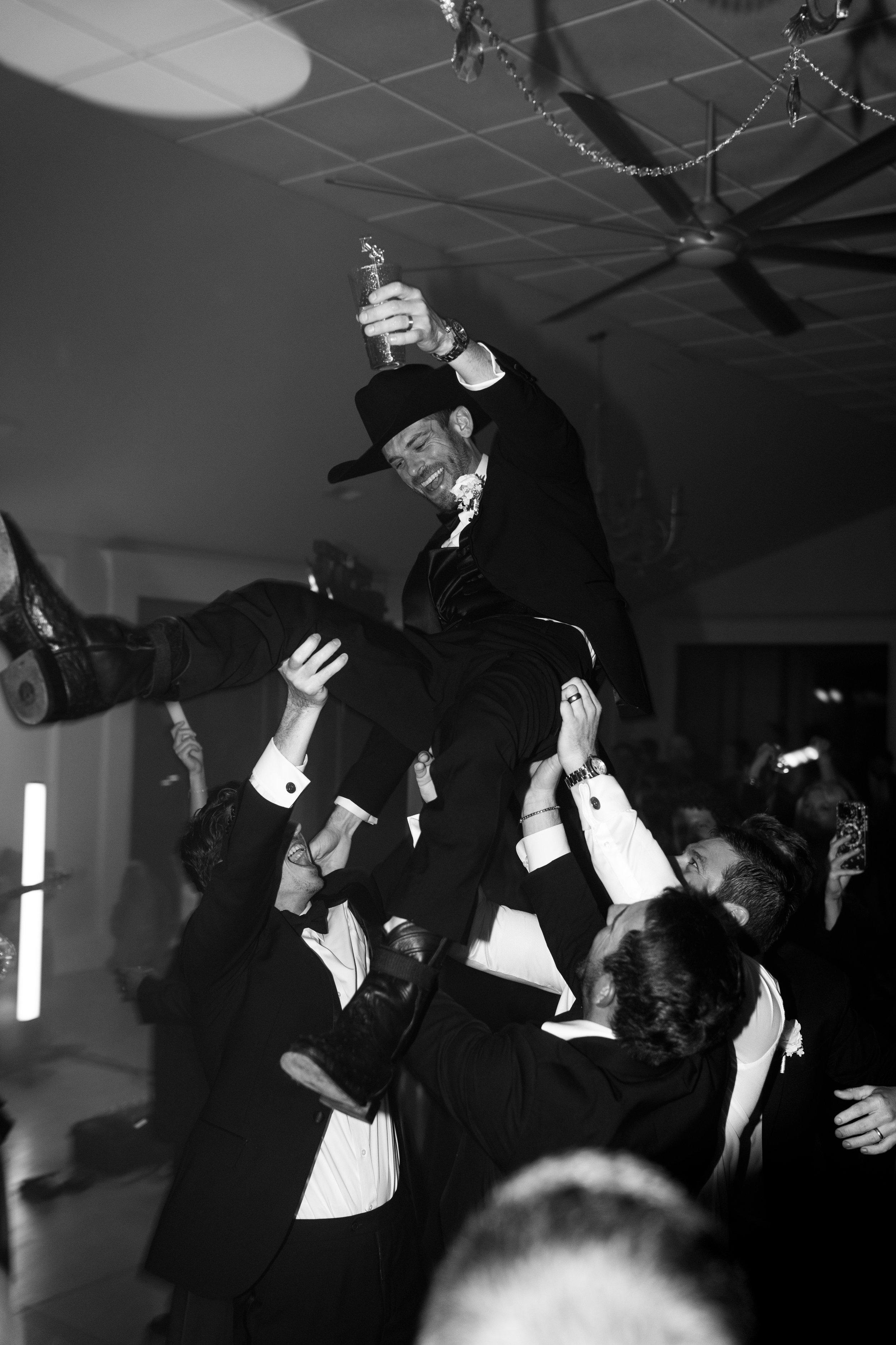 People at a celebration lifting a man in a suit and hat in the air, with one person holding a drink, in a decorated room.