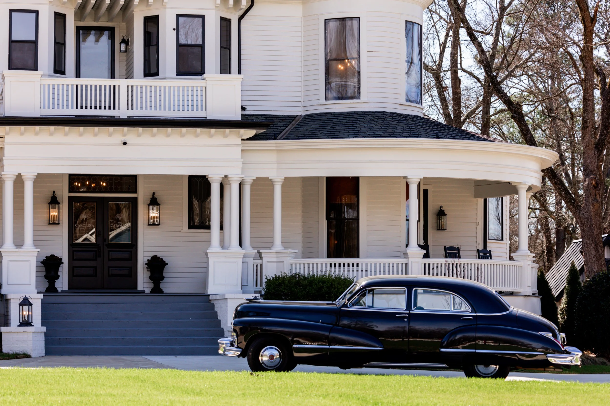 A vintage black car parked in front of a large white Victorian-style house with a wraparound porch and multiple windows, surrounded by trees.