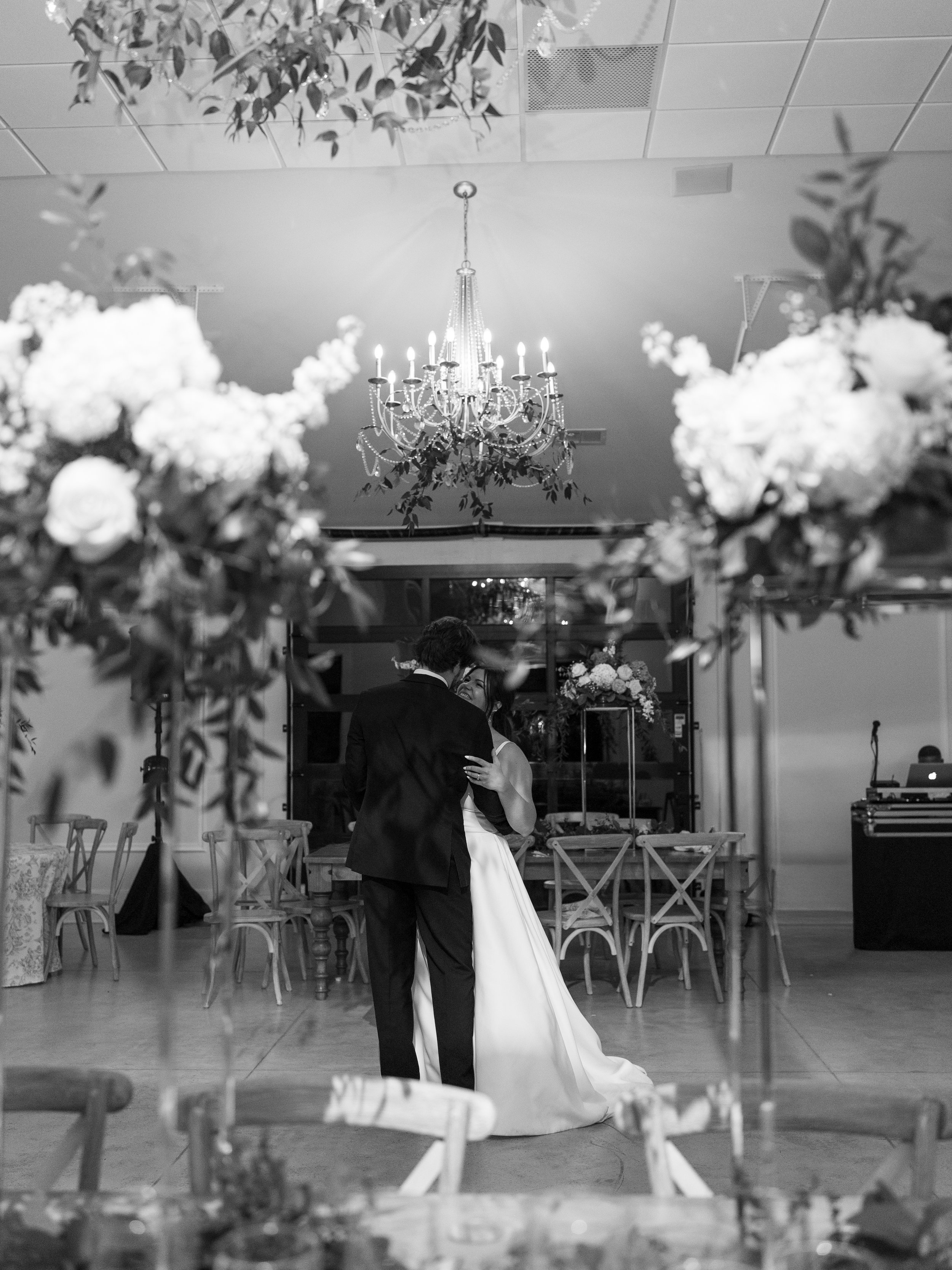 A black and white photograph of a bride and groom dancing at their wedding reception, with a chandelier hanging from the ceiling and floral decorations surrounding them.
