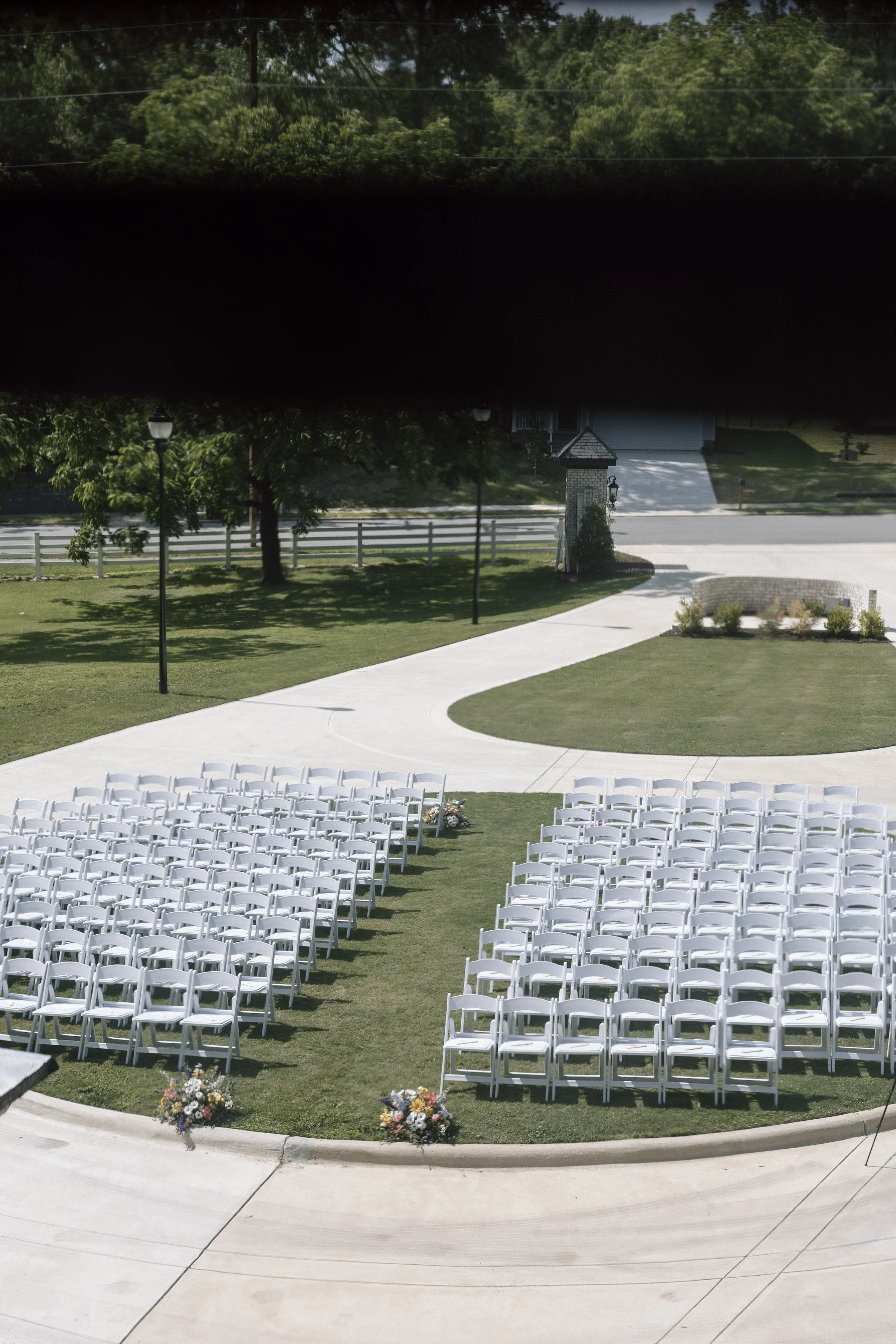 Wedding ceremony setup with rows of white chairs and floral arrangements on the grass, outdoor area with trees, lamp posts, and a pathway.