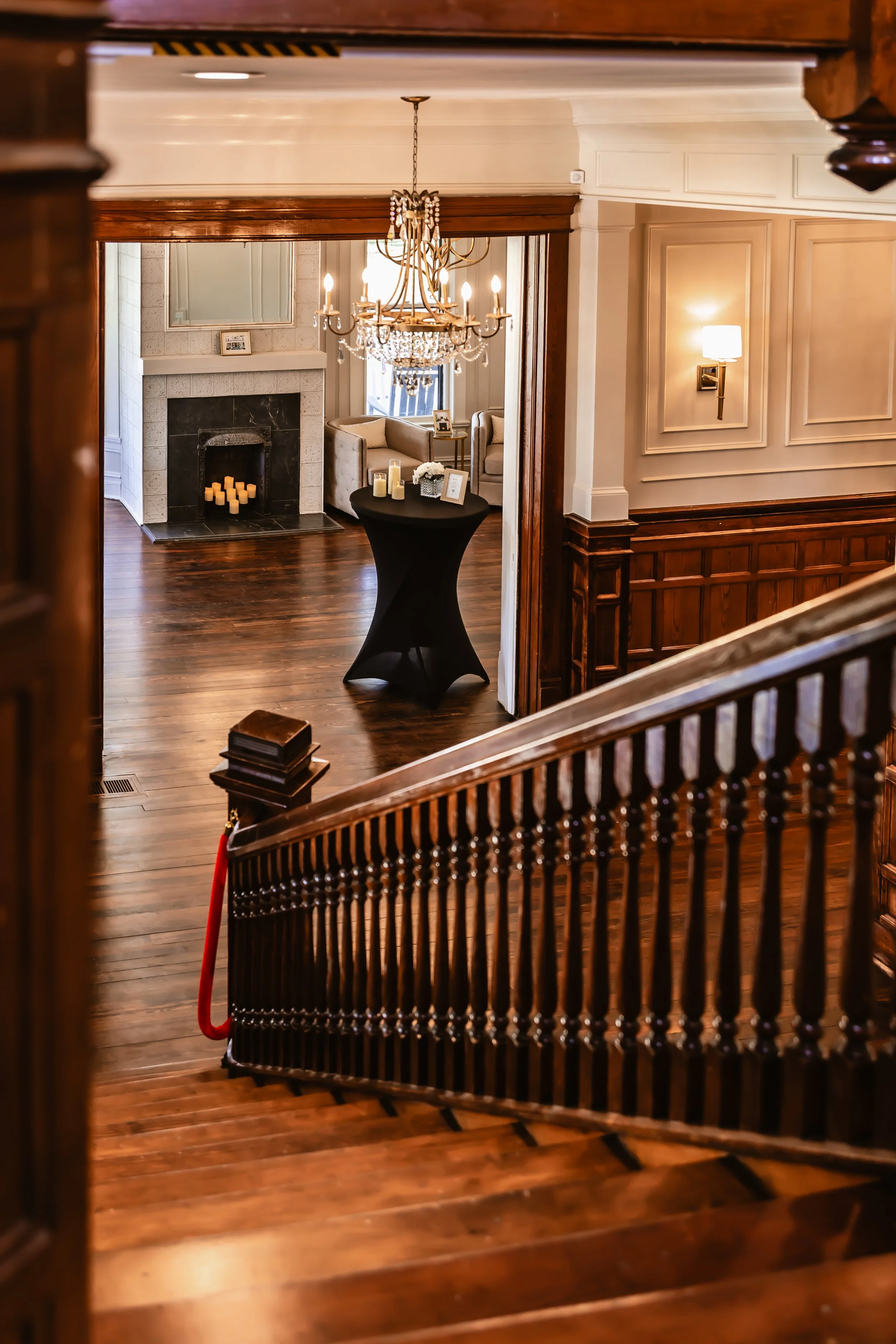 Elegant interior of a house with a staircase, hardwood floors, a chandelier, a fireplace with candles, and a sitting area with armchairs.