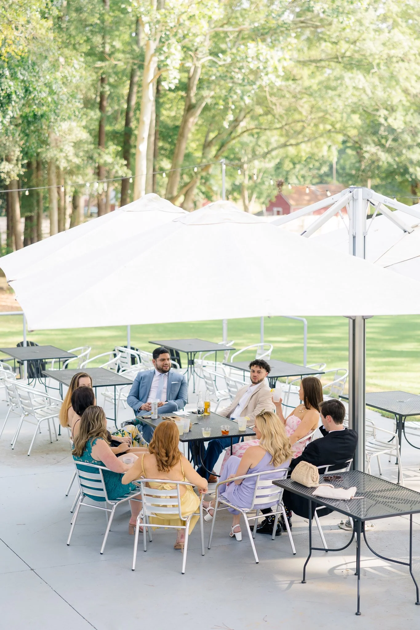 People socializing at a daytime outdoor gathering under large patio umbrellas, seated at tables with drinks in a lush green park-like setting.