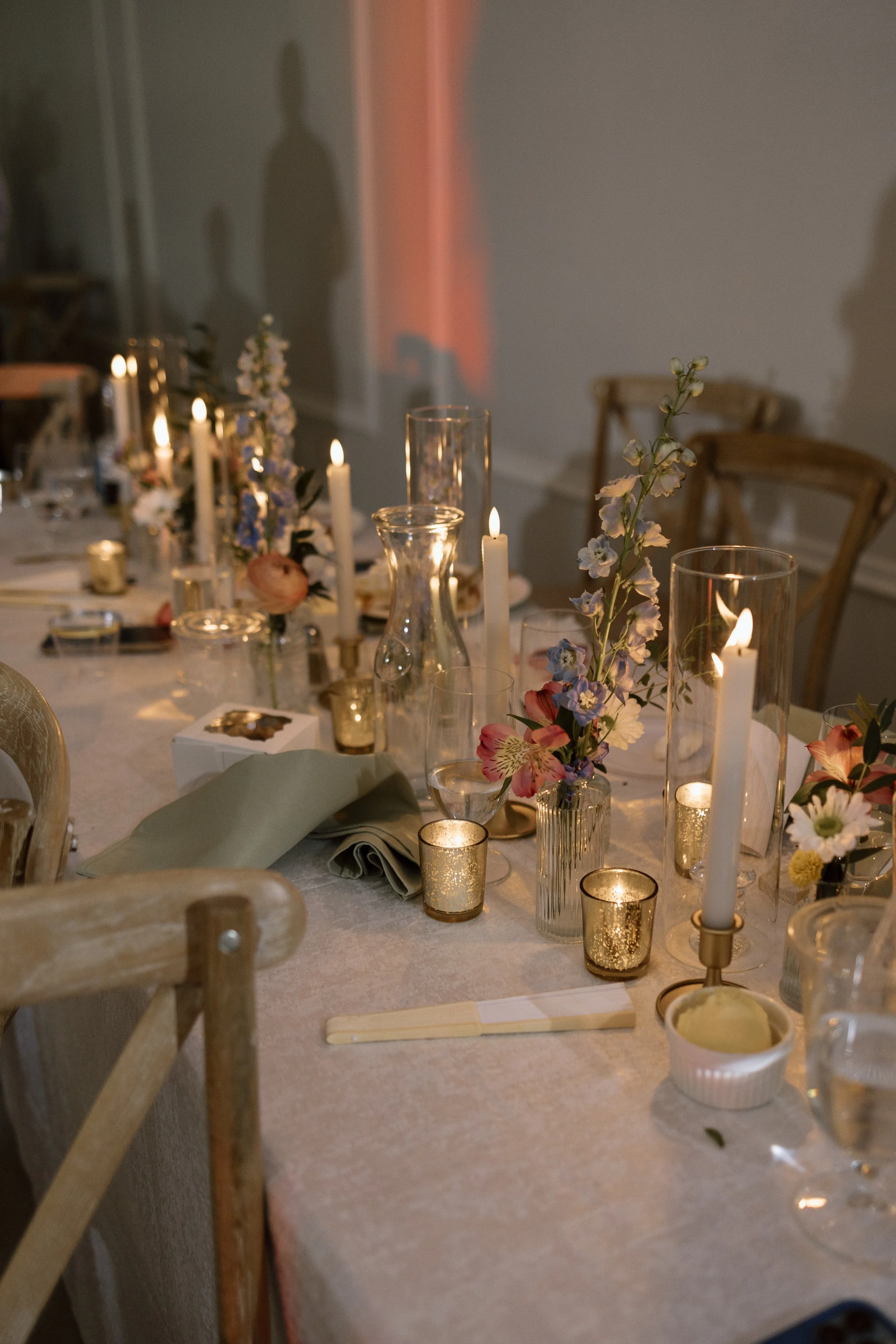 Decorated dining table with lit candles, floral arrangements, glasses, and napkins for a celebration or event.