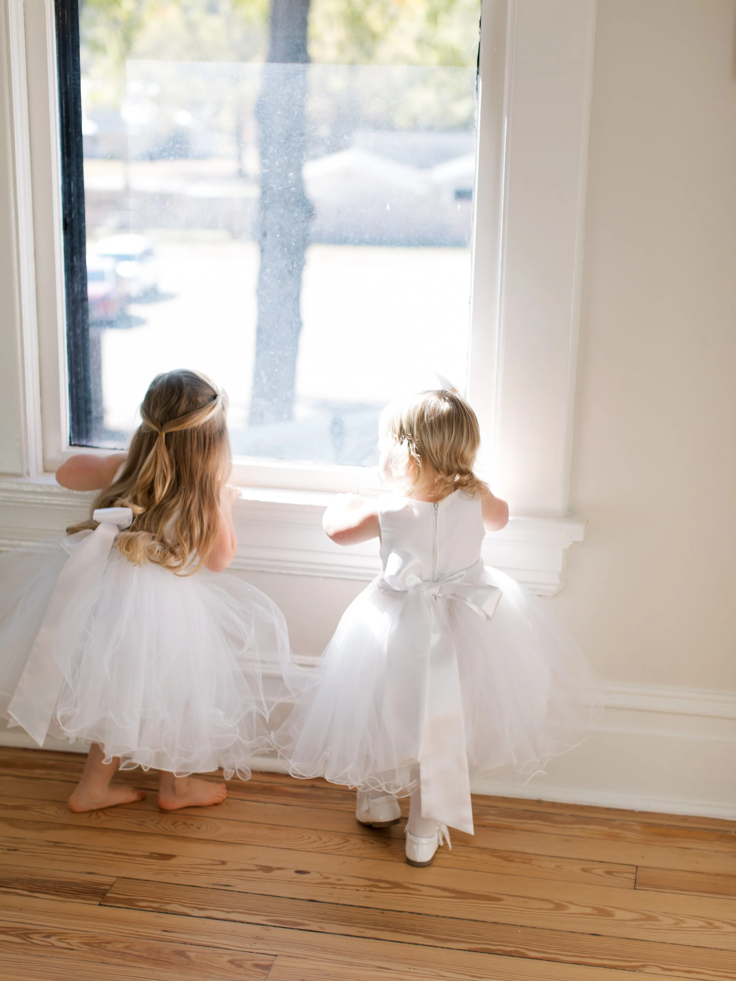 Two young girls in white dress looking out a large window.