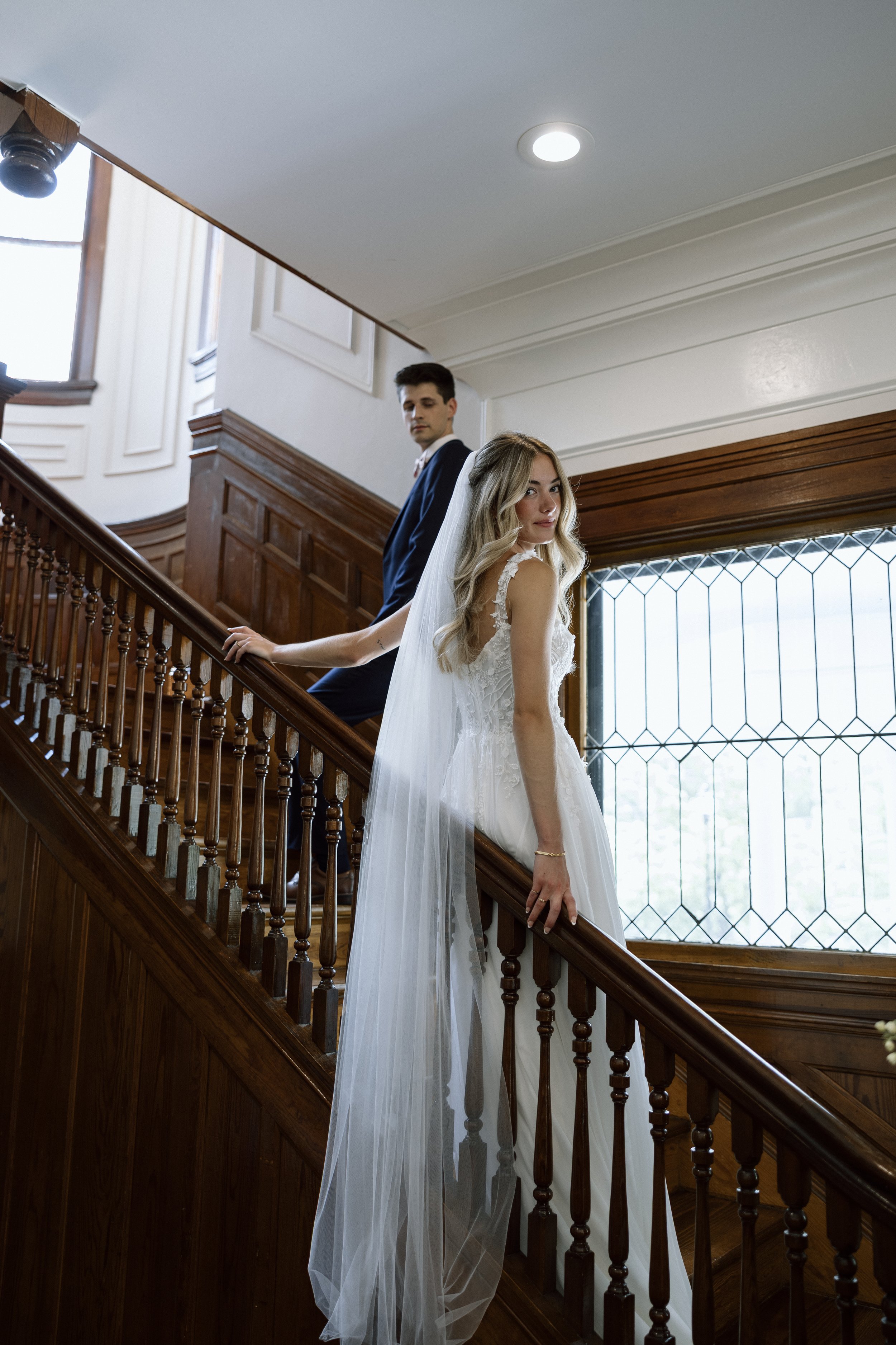 A bride and groom standing on a wooden staircase inside a building, with the bride in a white wedding gown and veil, and the groom in a dark suit, looking in different directions.