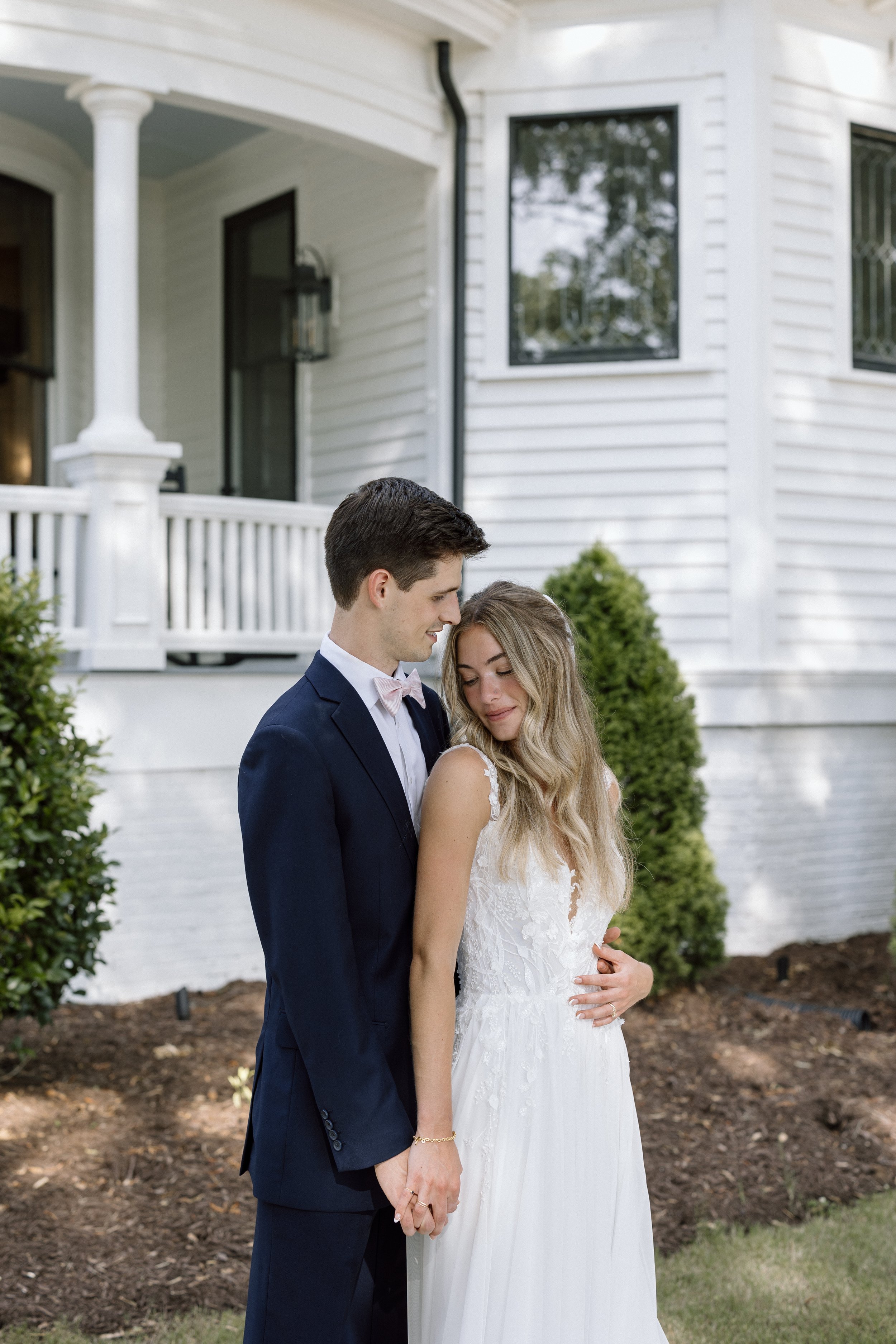 A wedding couple standing outside in front of a white house, holding hands and looking at each other, with greenery and soil in the background.
