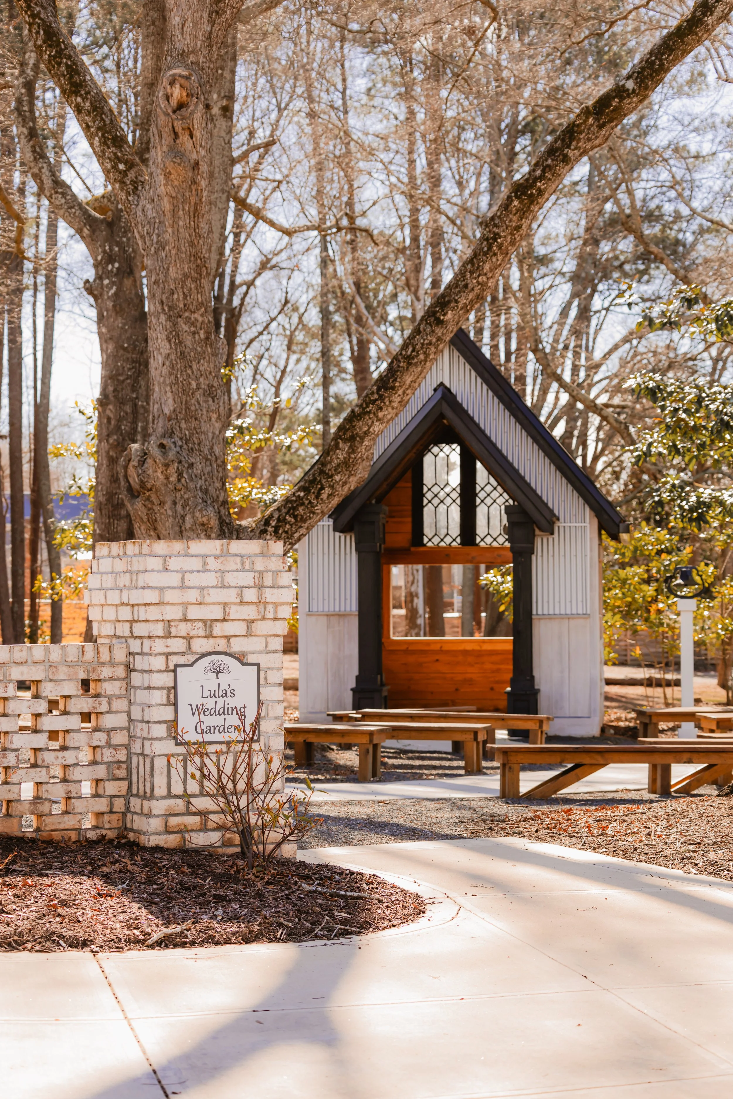 A small white chapel with black trim, set among trees with fall foliage, is located behind a brick wall with a sign that reads "Lula's Wedding Garden." Wooden benches are arranged in front of the chapel.