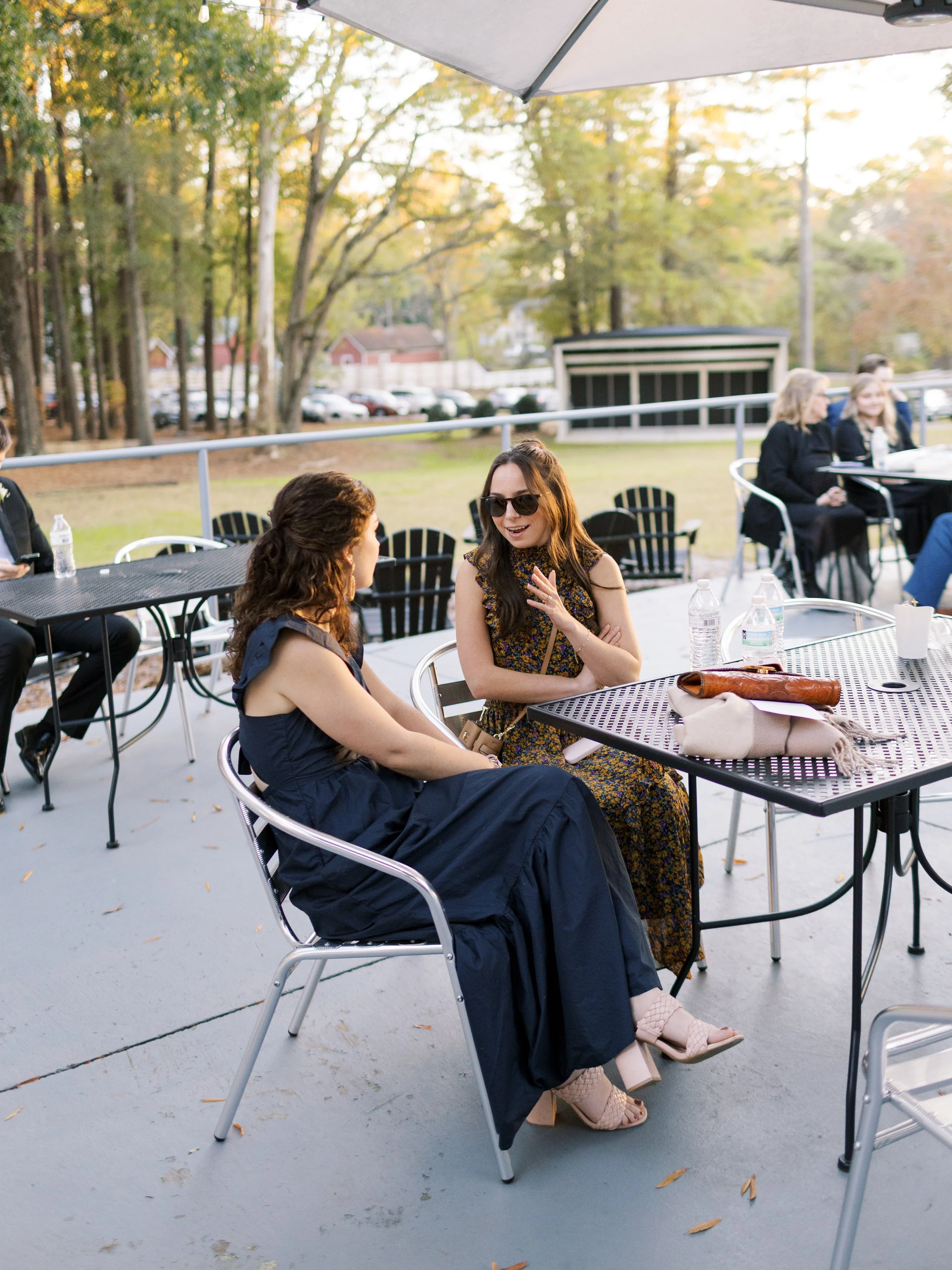 Two women engaged in conversation at an outdoor event, seated at a table with water bottles and a handbag, with other people in the background and trees with fall foliage.
