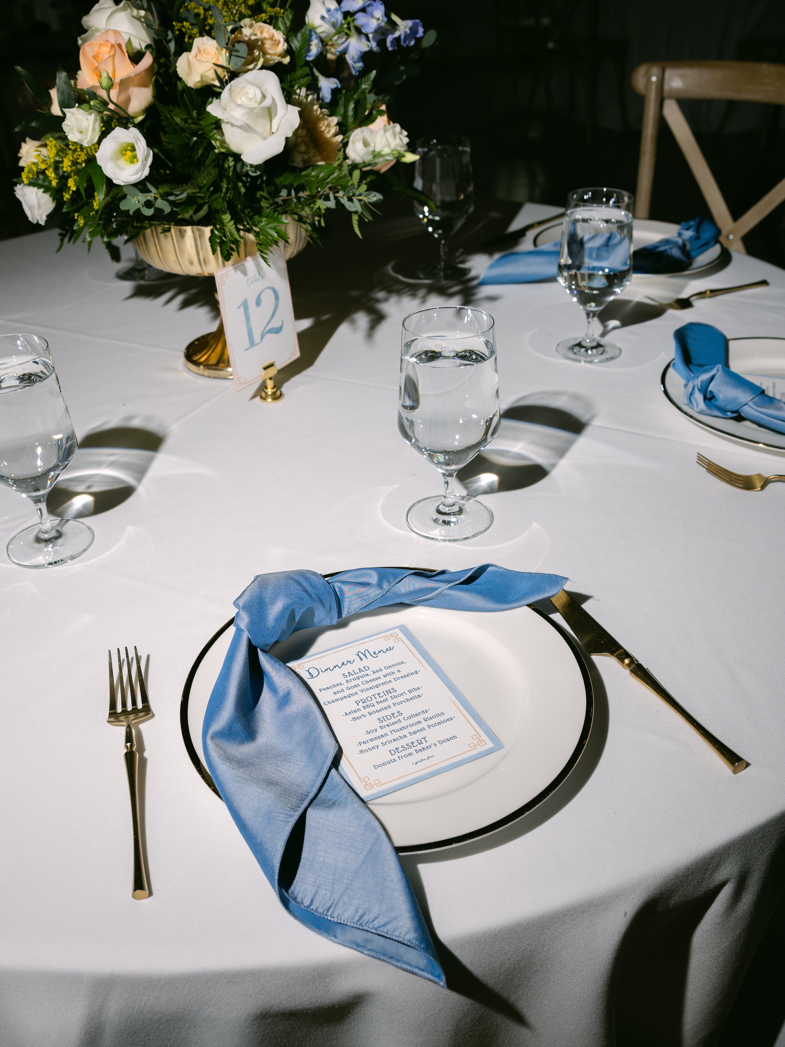A table set for a formal occasion with a white tablecloth, a floral centerpiece, a place setting with a white plate, gold flatware, a blue cloth napkin, and a dinner menu. Water glasses are filled, and a table number card is in the center.