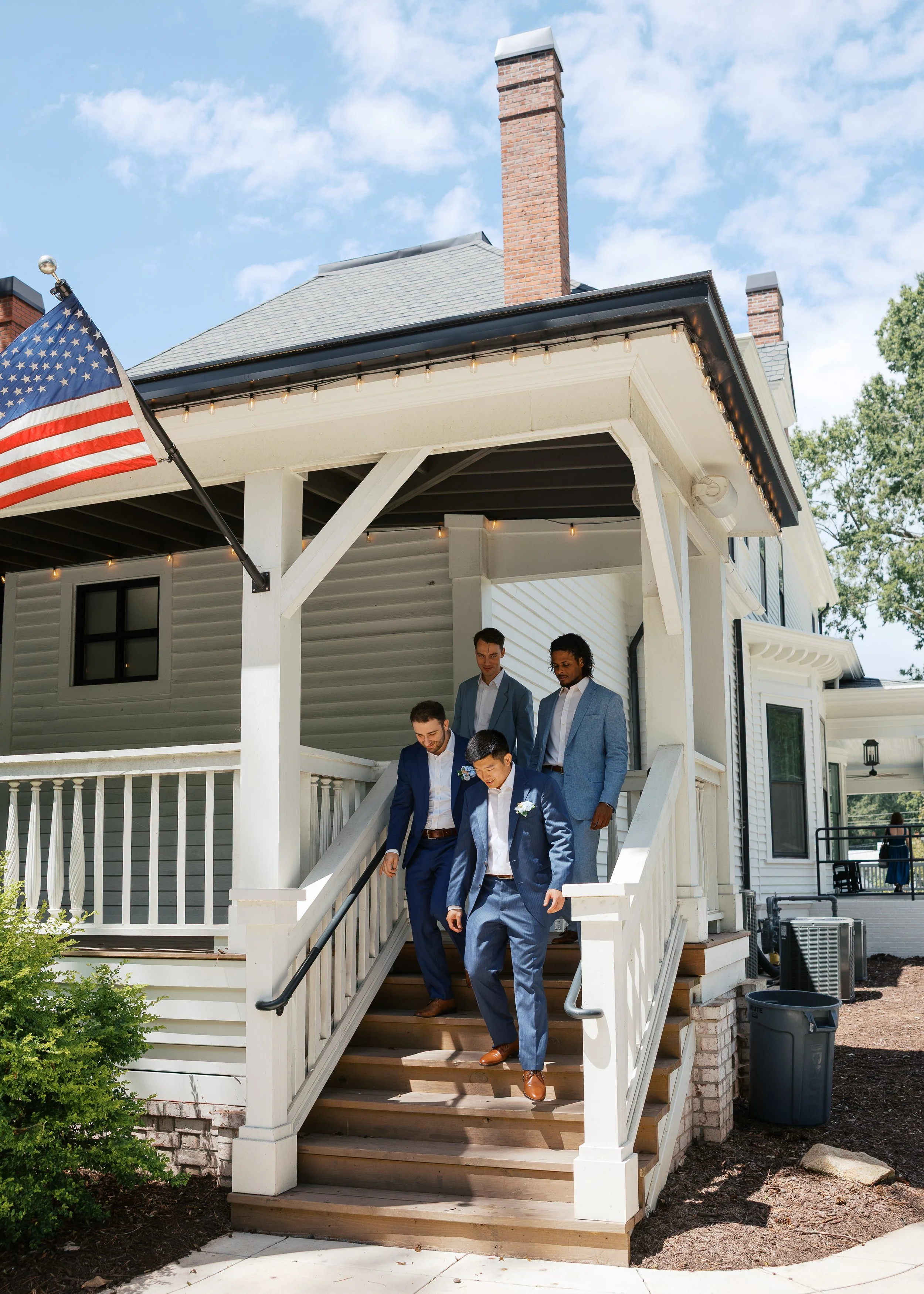 A group of men in suits descending the front steps of a house during daytime, with an American flag hanging nearby.