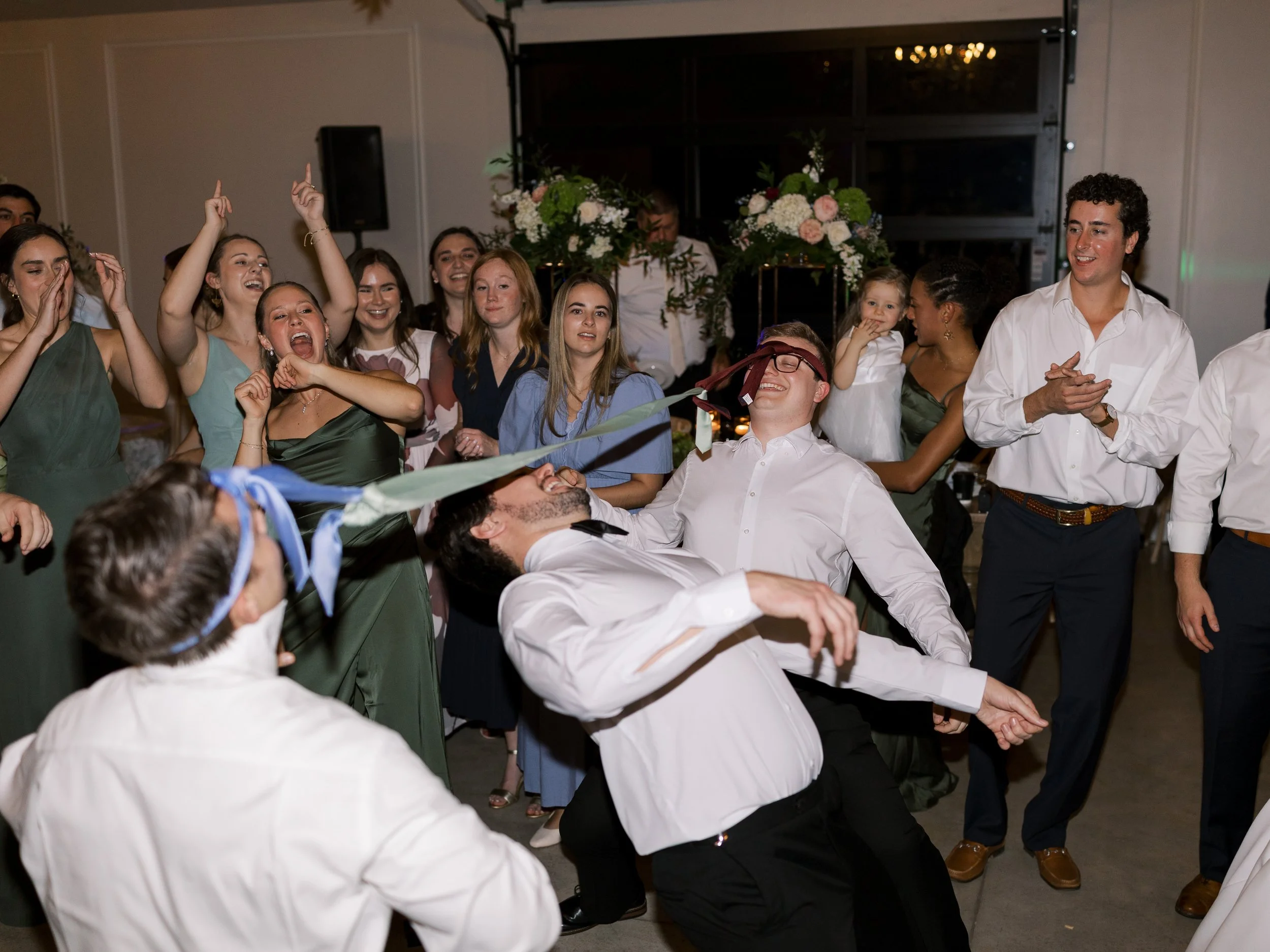 Group of people at a celebration, with two men in white shirts pulling on a ribbon during a game or activity, surrounded by friends clapping and cheering, decorated with floral arrangements.