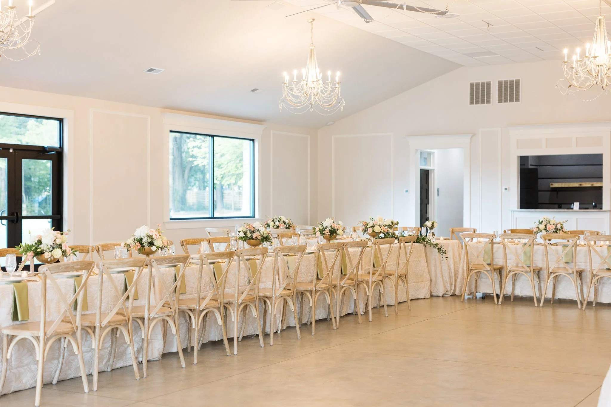 Wedding reception room with long banquet tables decorated with floral centerpieces and surrounded by wooden cross-back chairs, chandeliers hanging from the ceiling, large windows letting in natural light, and a wooden bar area in the background.