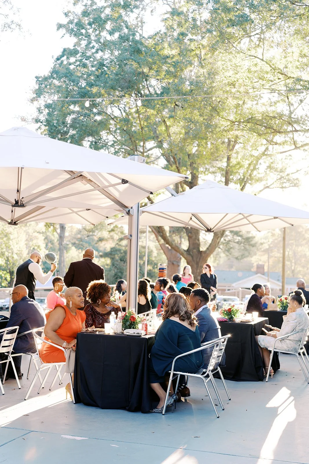 People sitting at outdoor tables under umbrellas at a social event in a park with large trees in the background.