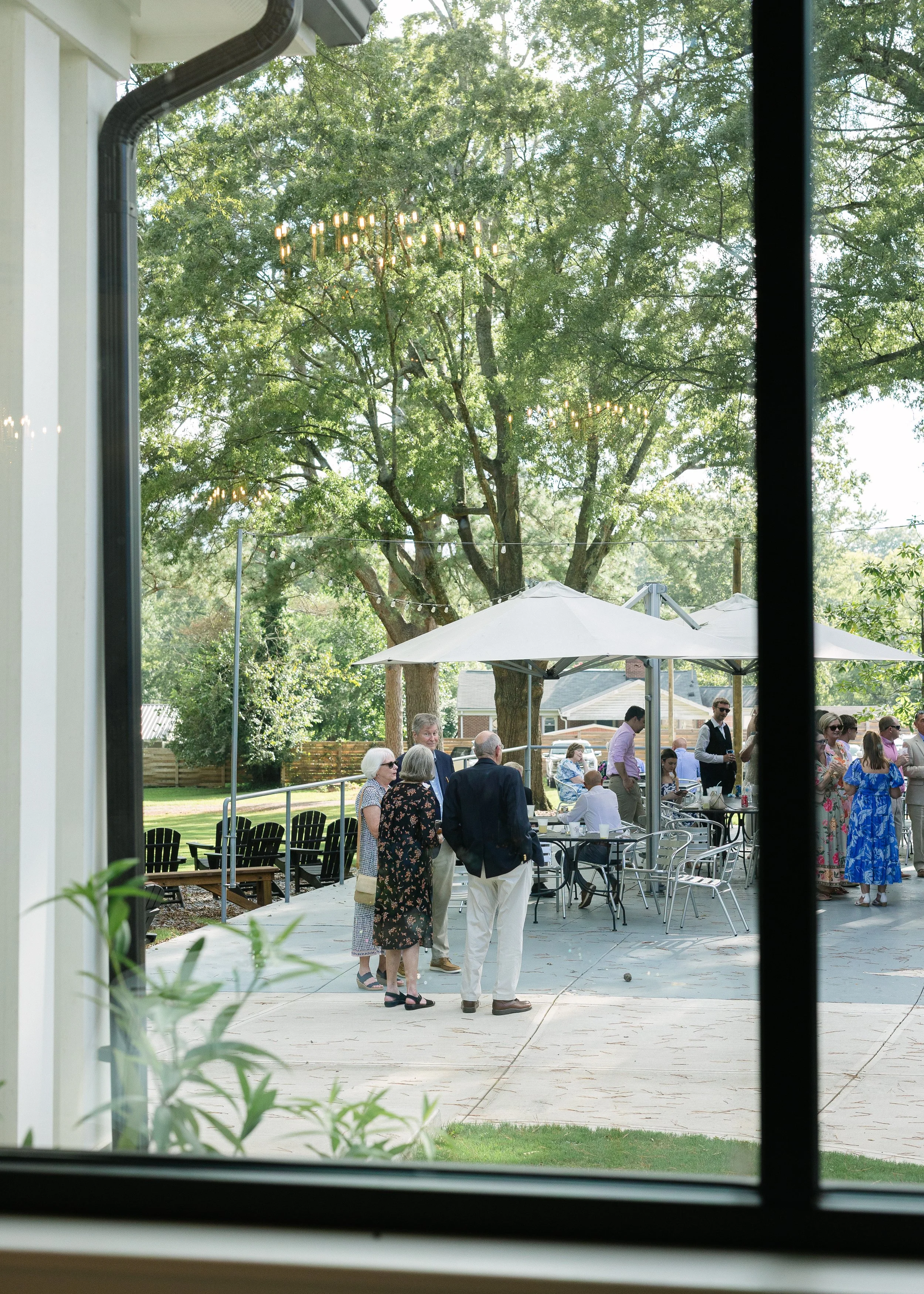 People gathered in a backyard patio during a social event, with some standing and talking, others sitting under large umbrellas, with trees and a chandelier visible in the background.