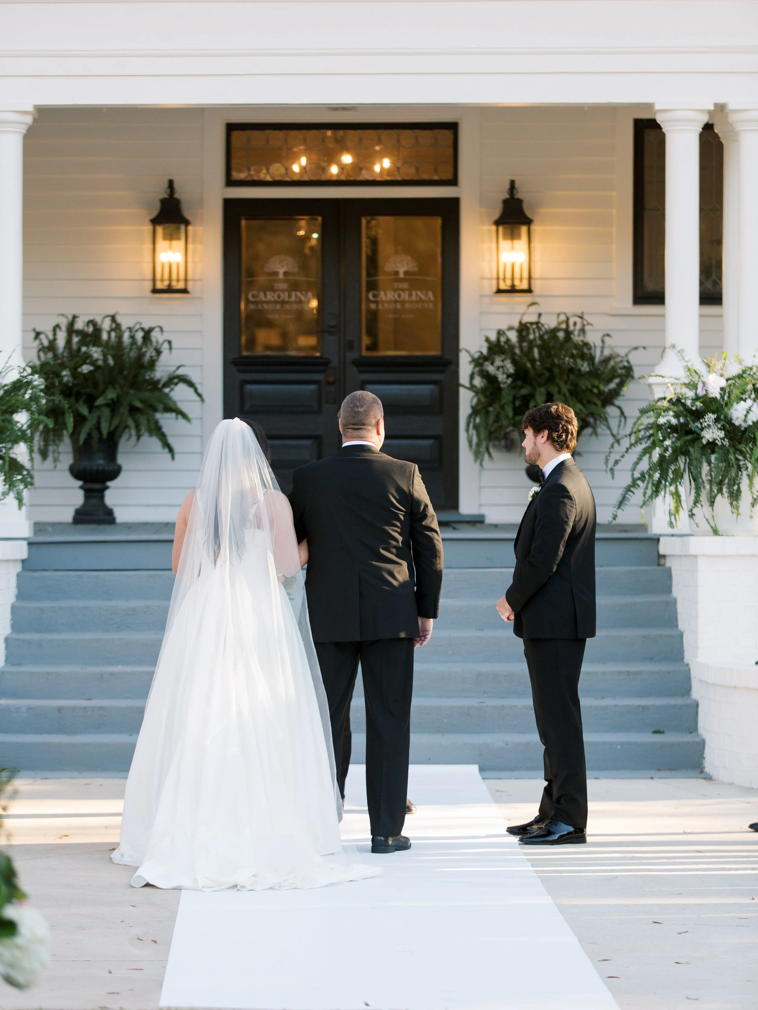 Bride and groom standing with officiant outside a white house with black door, preparing for wedding ceremony.