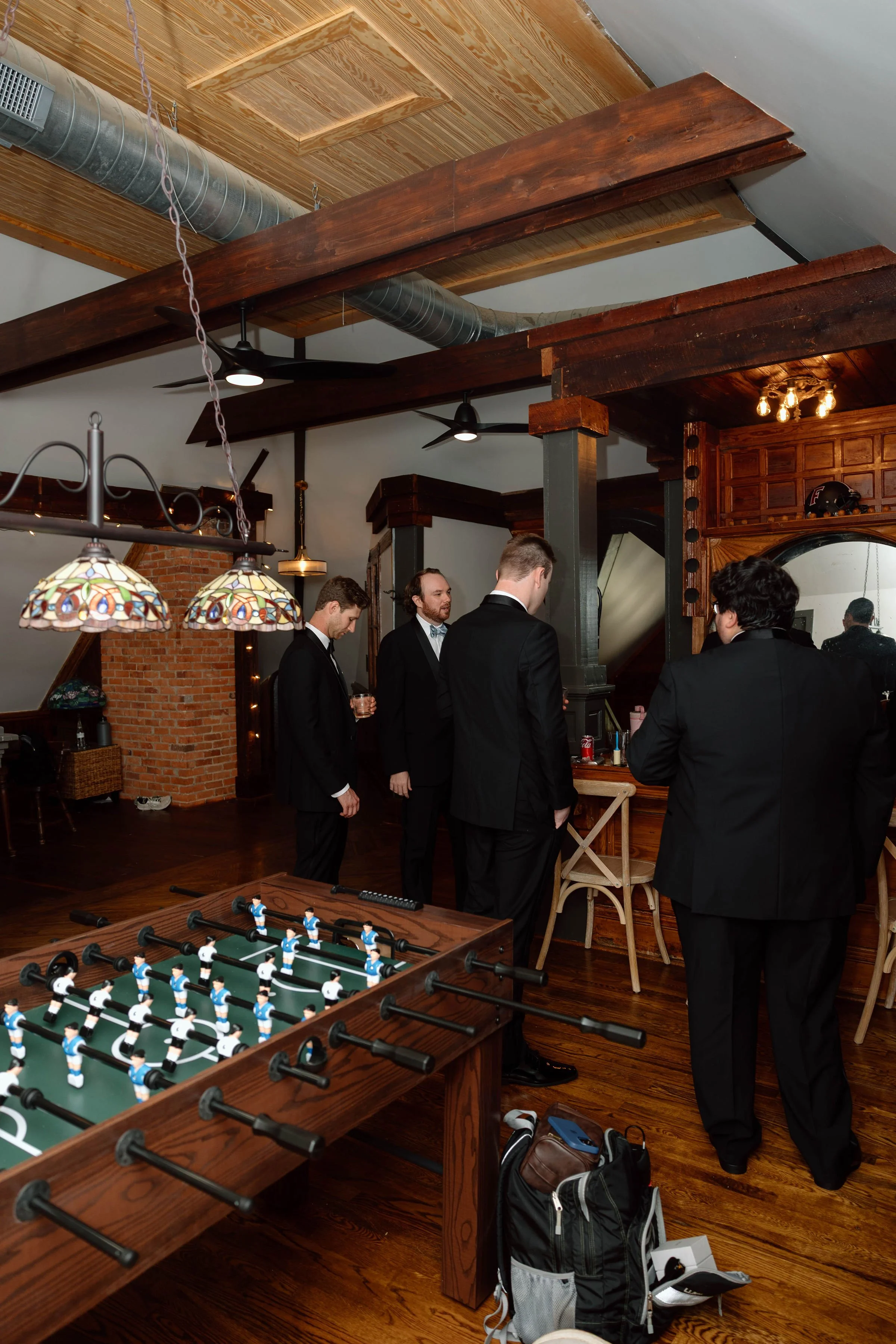 A group of men in formal suits gathered in a room with wood-paneled walls, playing a game of foosball. The room features stained glass pendant lights, exposed brick, wooden beams, and a foosball table.