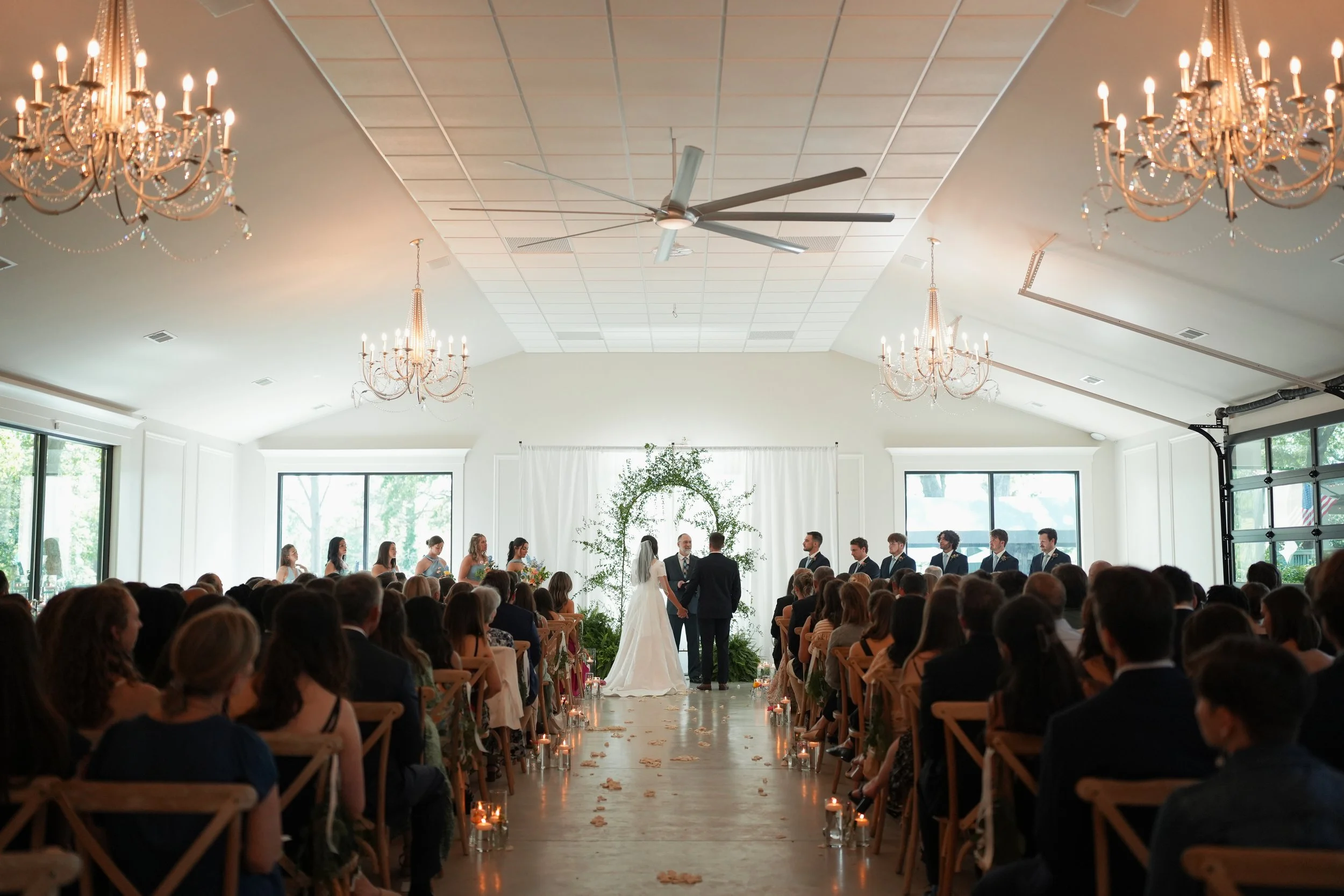 Indoor wedding ceremony with bride and groom standing under floral arch, surrounded by seated guests, bridesmaids and groomsmen on either side, candles along aisle, chandeliers hanging from ceiling.