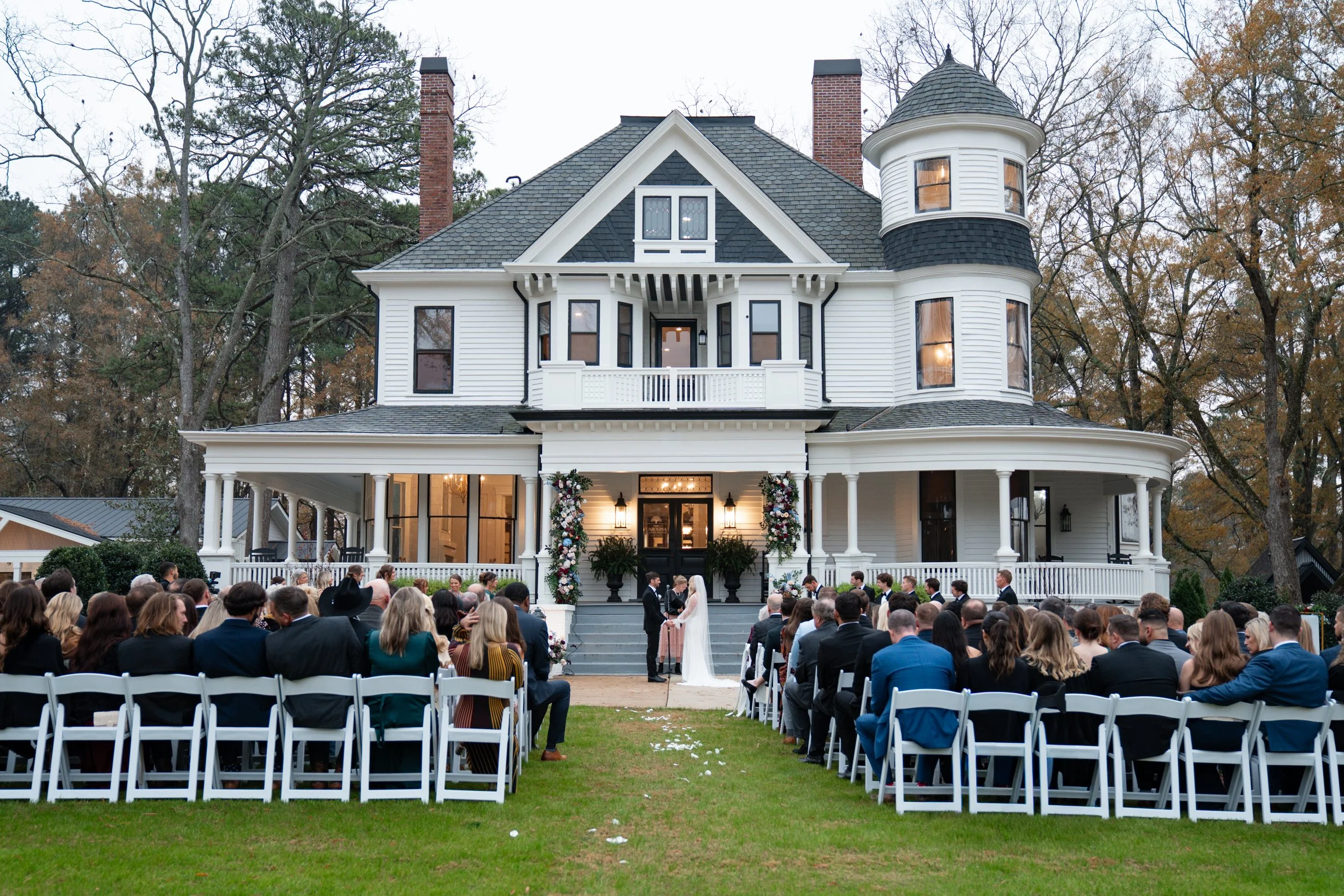 A wedding ceremony taking place outdoors in front of a large, historic white house with multiple stories and a tower. Guests are seated in white chairs facing the couple at the altar.