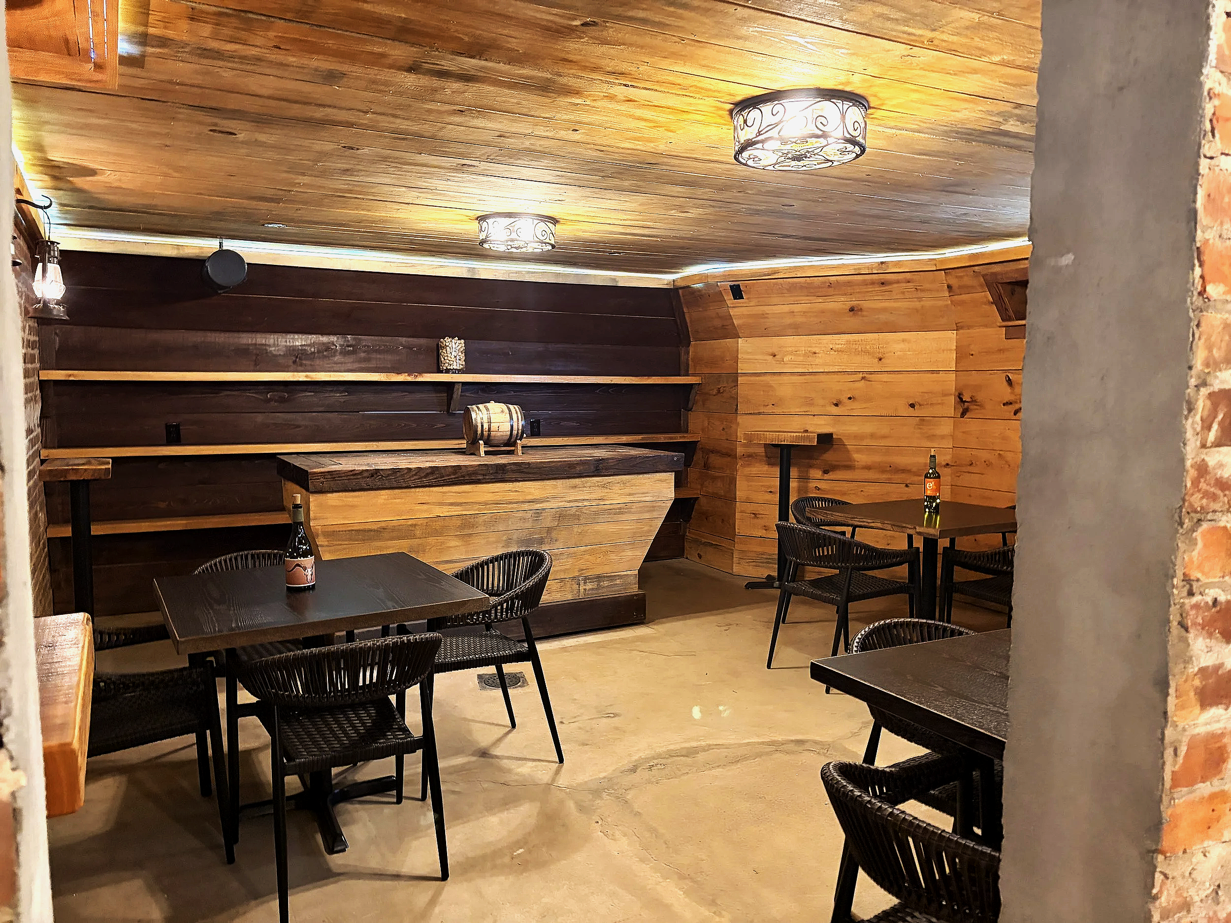 Interior of a rustic room with a brick wall displaying the logo of The Carolina Manor House, featuring a stylized tree. The room is lit by vintage-style hanging lanterns and modern ceiling lights. There is a wooden bar or counter with shelving behind it, and the ceiling and part of the wall are covered in wood paneling.