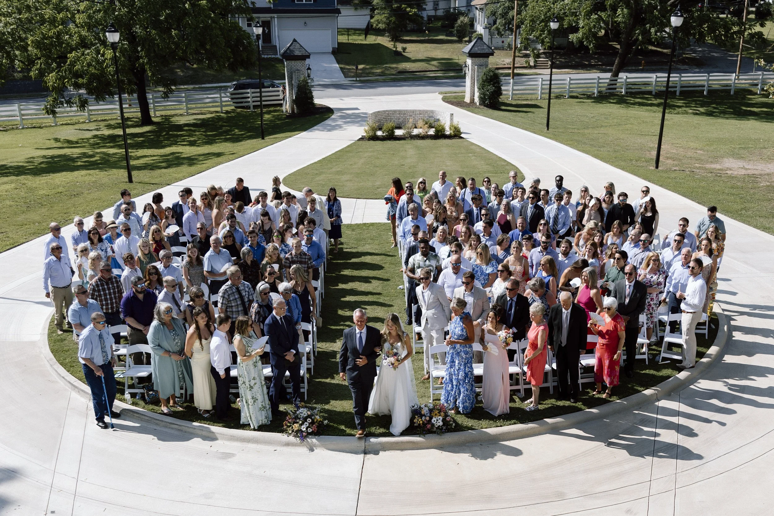An outdoor wedding ceremony with numerous guests seated on white chairs on a grassy area, with a bride and groom walking down the aisle surrounded by family and friends.