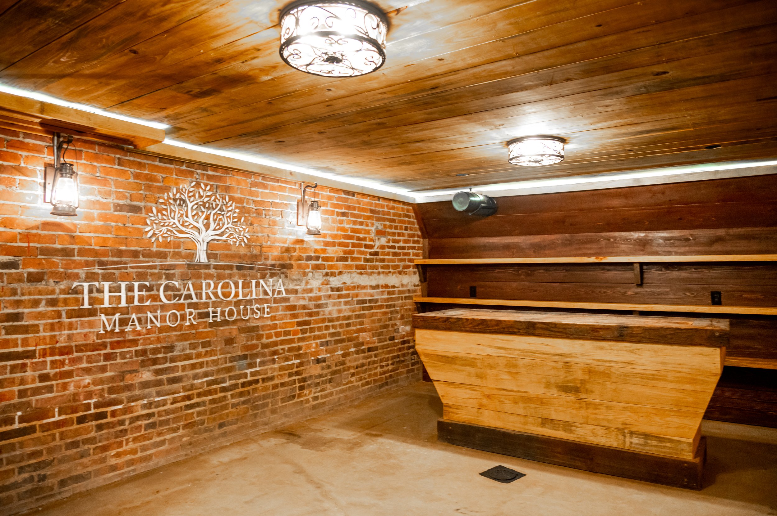 Interior of The Carolina Manor House with a brick wall, wooden shelves, decorative lighting, and a sign with a tree logo.